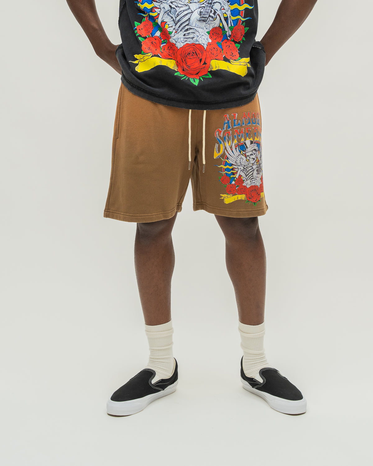 A person wearing tan drawstring shorts with a colorful skeleton graphic on the left leg, paired with a black graphic t-shirt, white socks, and black slip-on shoes, photographed against a plain light background.