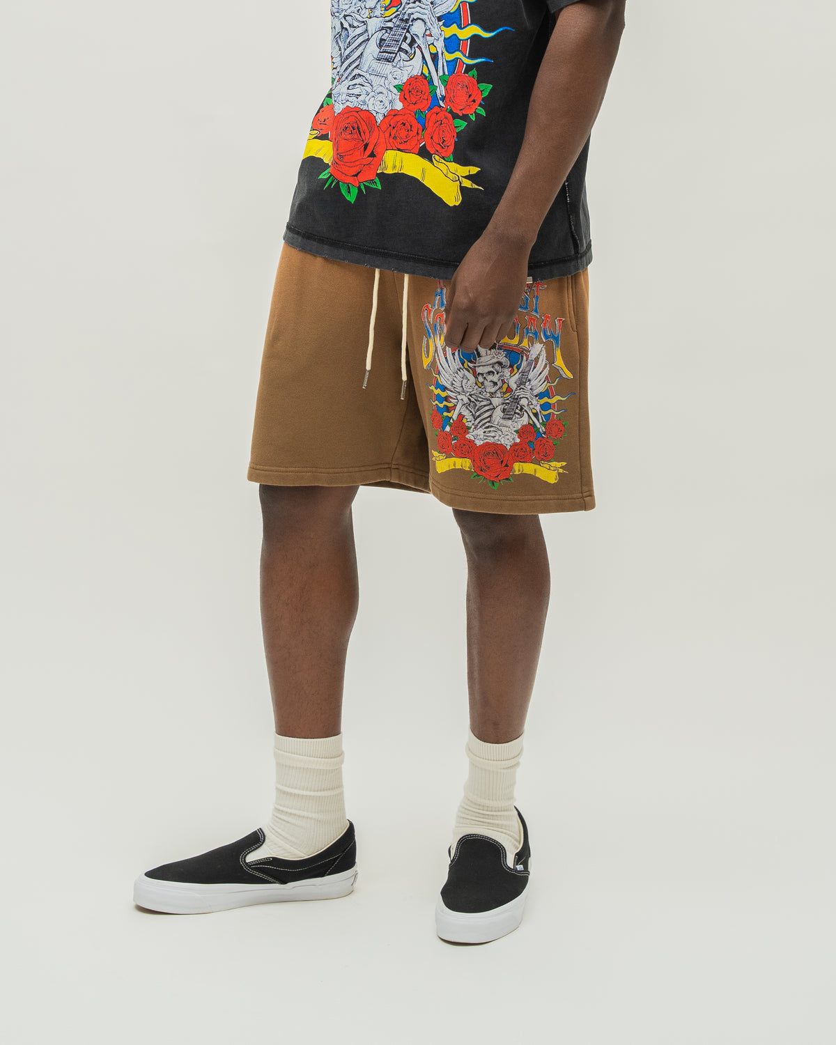 A model wearing brown drawstring shorts with colorful skeleton and floral graphics on the front, paired with a black graphic t-shirt, white socks, and black slip-on sneakers, photographed against a light gray studio background.