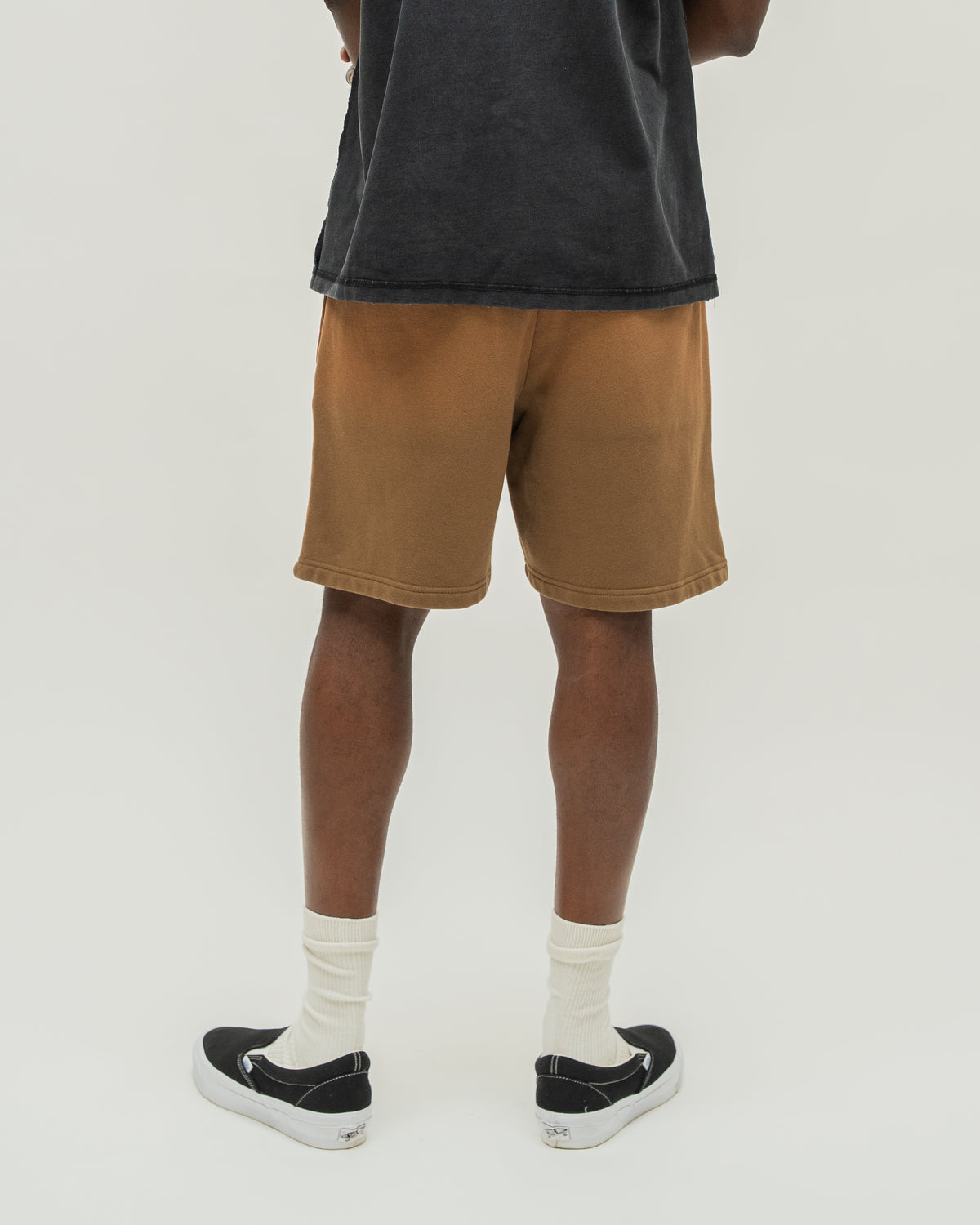 A person shown from the back wearing brown washed denim shorts, a dark charcoal t-shirt, white socks, and black slip-on sneakers against a light gray studio background.