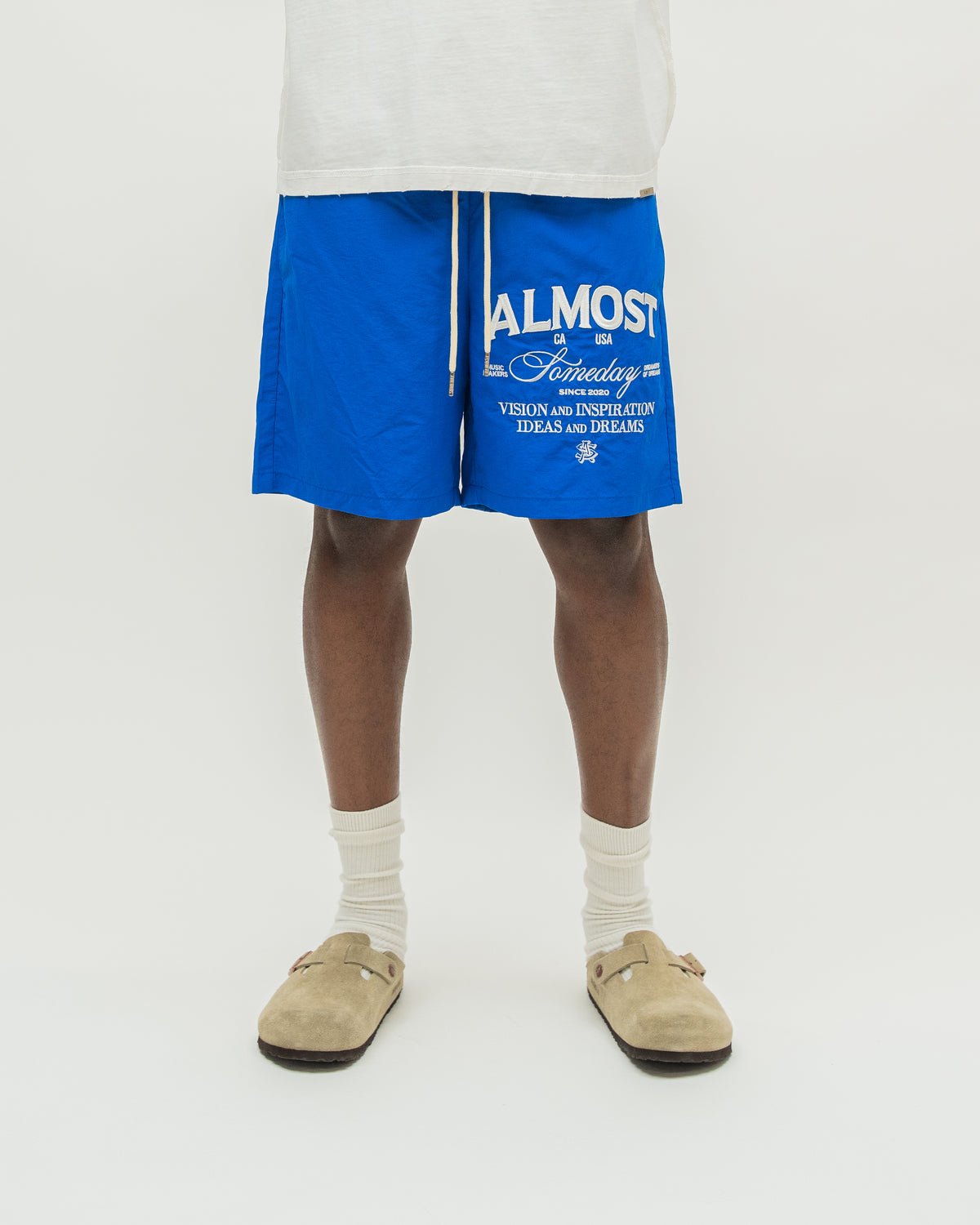 A person from the waist down wearing bright blue drawstring shorts with white text, white socks, and beige slip-on shoes against a plain light background.