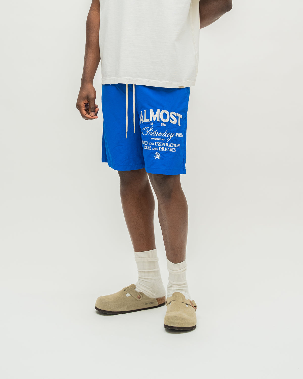 A model from the waist down wearing bright blue drawstring shorts with white graphic text, a white t-shirt, white socks, and beige clogs, photographed against a clean white studio background.