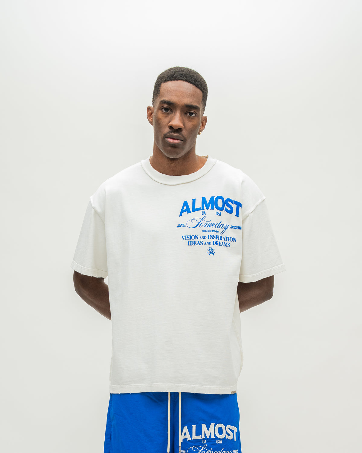 A man modeling an oversized off-white t-shirt with blue graphic text on the chest that reads 'ALMOST SOMEDAY, paired with blue shorts against a plain light gray background.