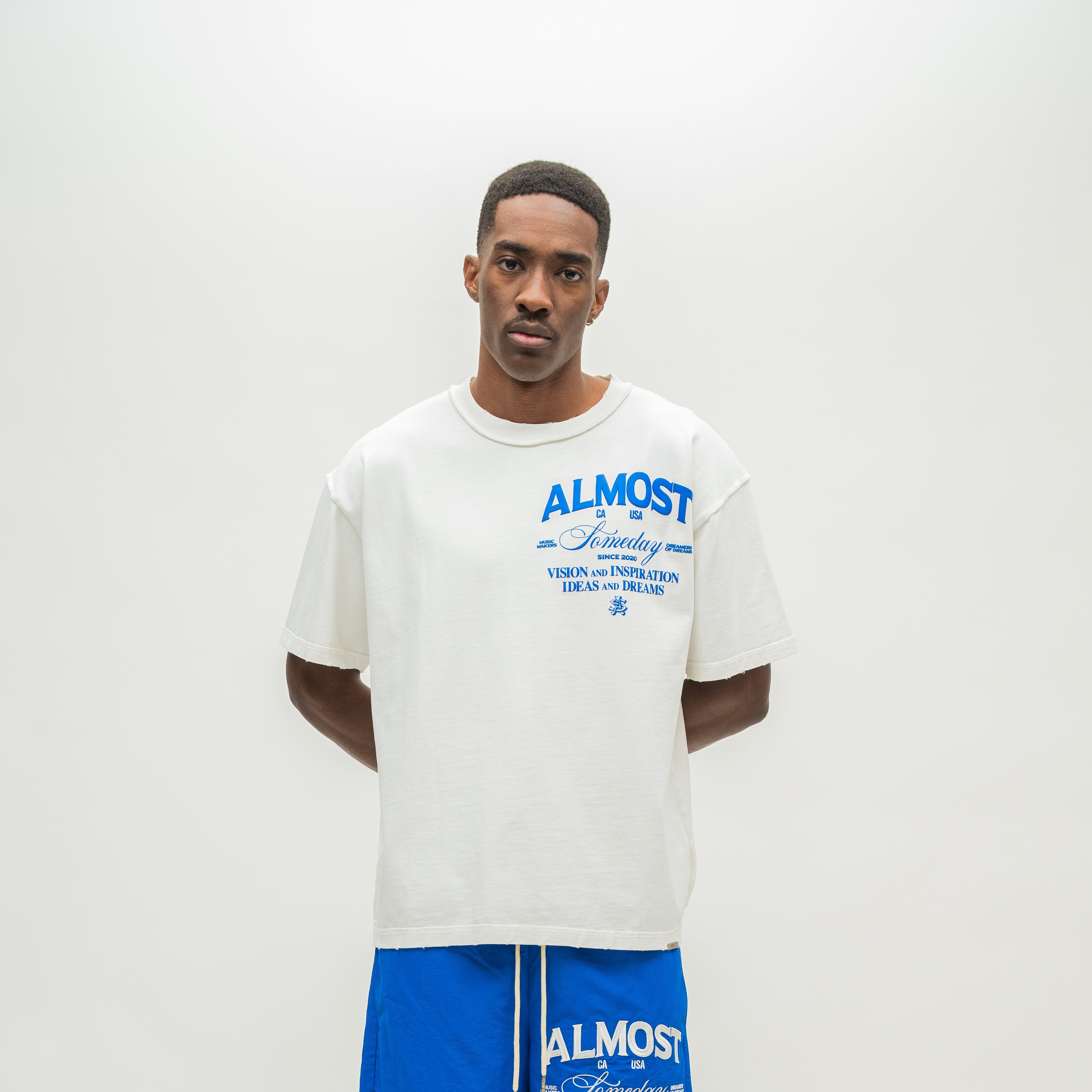 A man modeling an oversized off-white t-shirt with blue graphic text on the chest that reads 'ALMOST SOMEDAY, paired with blue shorts against a plain light gray background.
