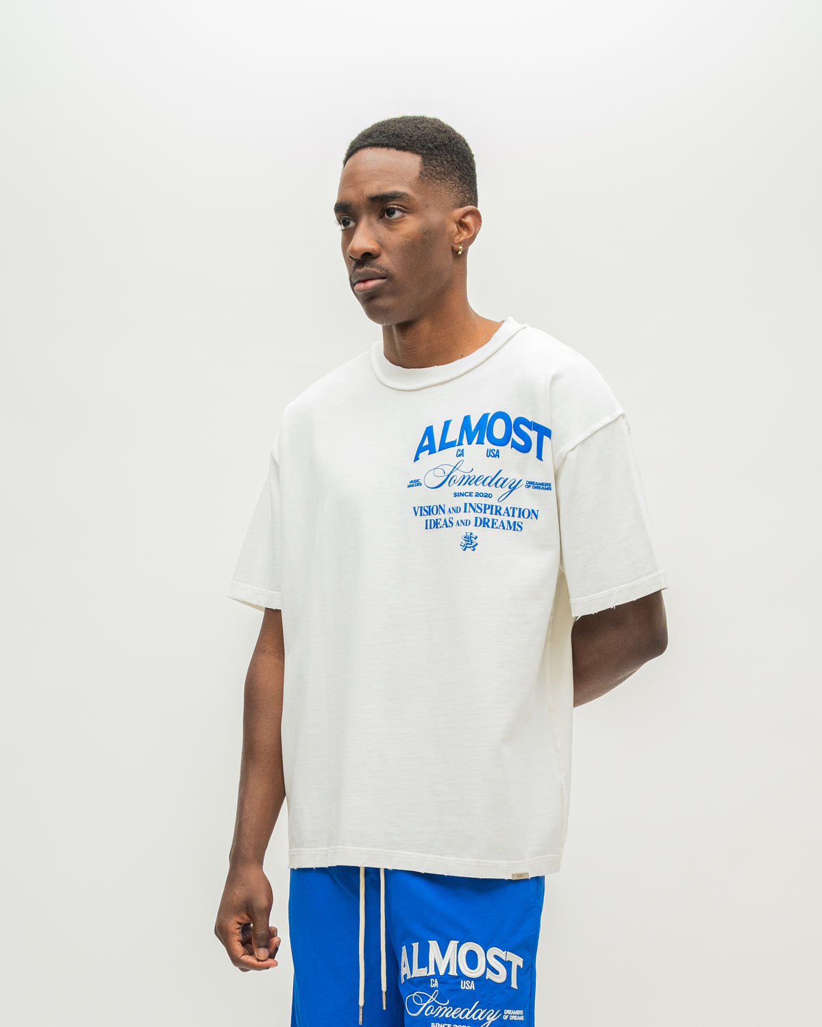 A man wearing an oversized white t-shirt with blue graphic text that reads 'ALMOST SOMEDAY' and blue athletic shorts with matching blue text, standing against a plain off-white studio background.