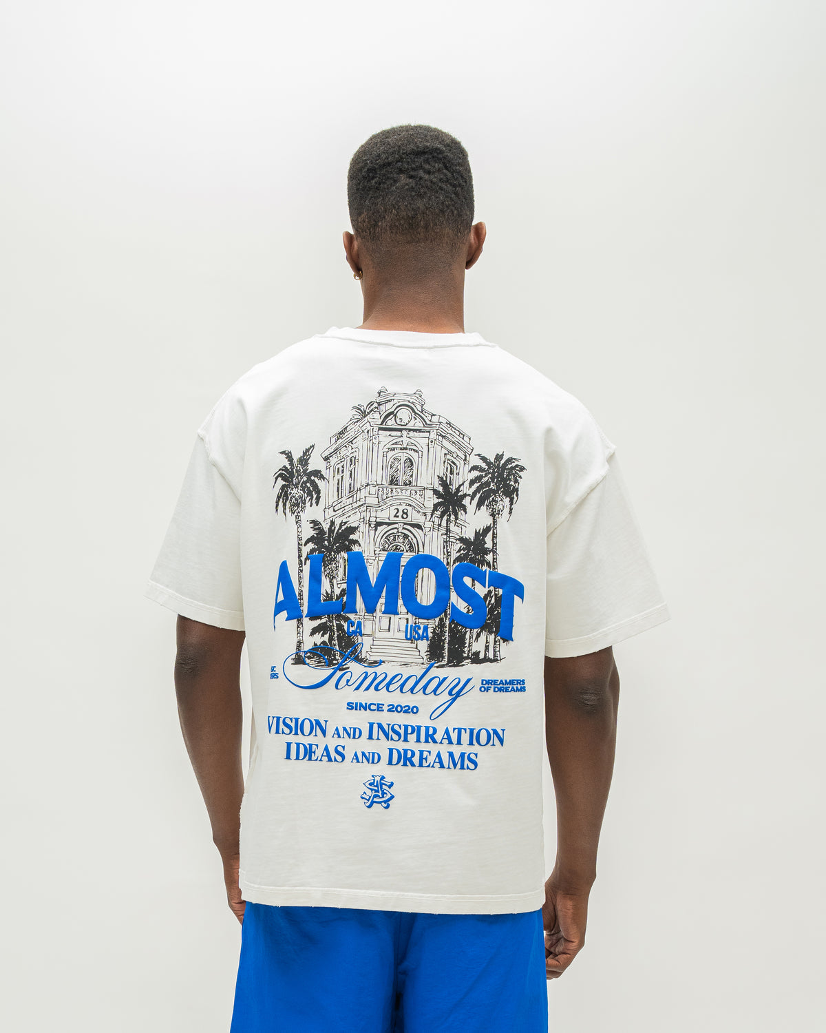 A man viewed from behind wearing an oversized off-white graphic t-shirt with a blue and black illustration and text on the back, paired with bright blue shorts, against a plain white studio background.
