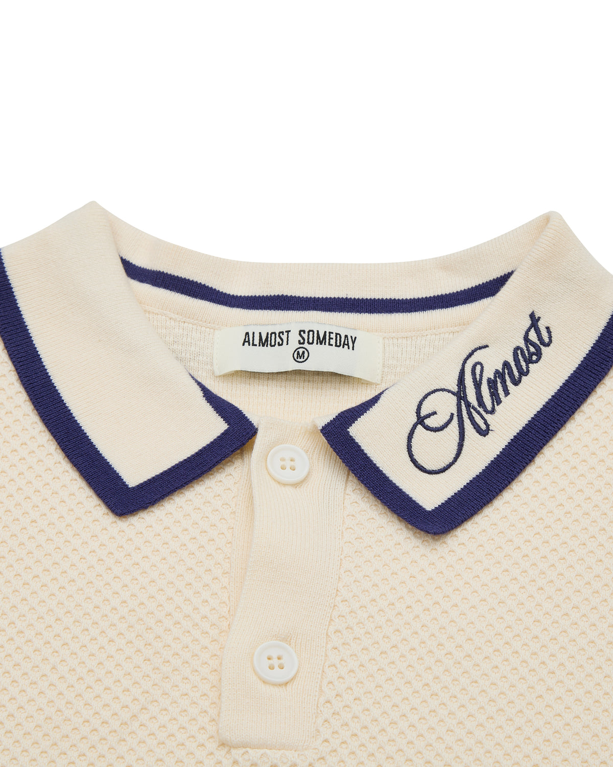 Close-up of a cream-colored polo shirt with navy trim and 'Almost' branding.
