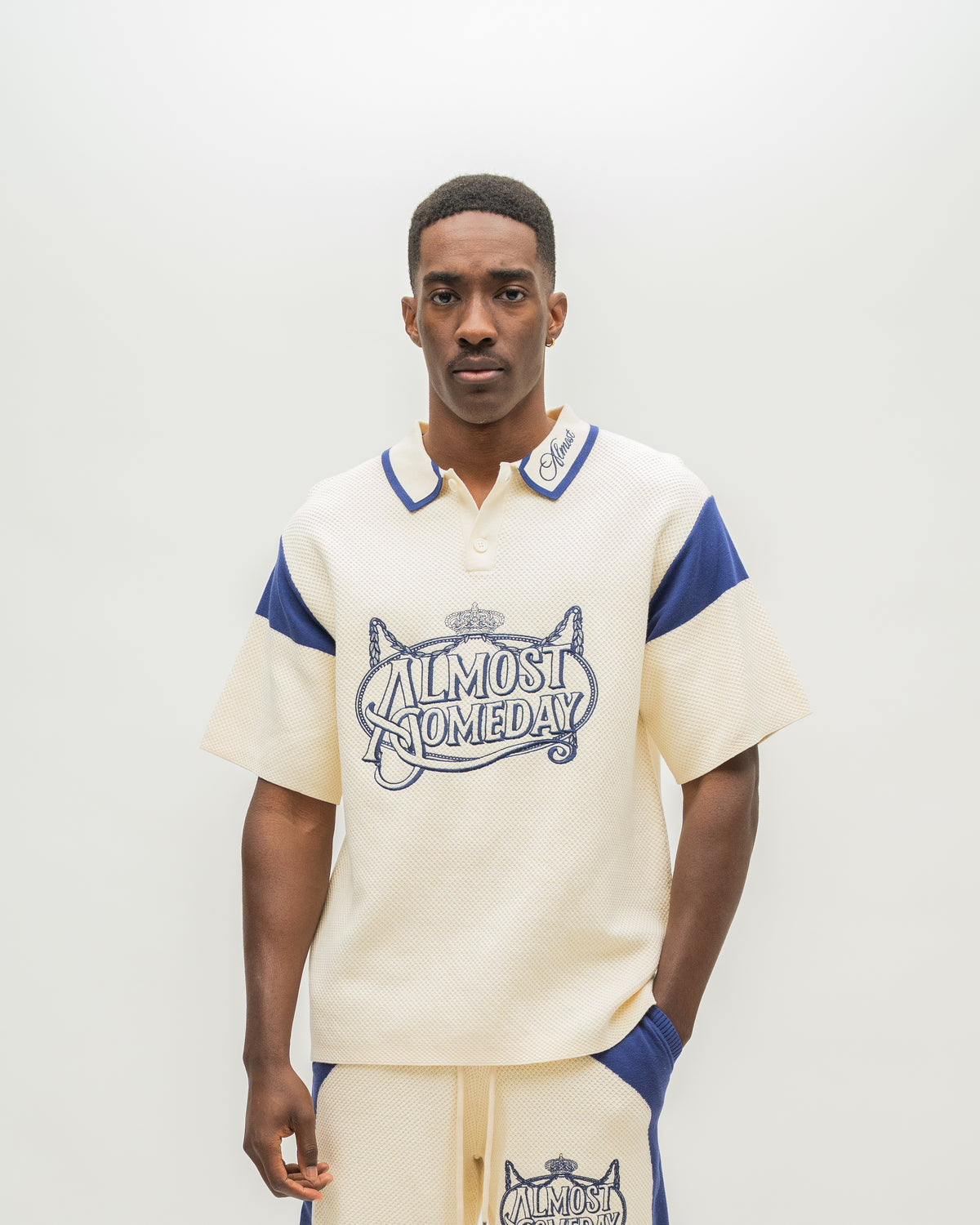 A man modeling a cream-colored short-sleeve polo shirt with blue accents and a graphic reading 'ALMOST SOMEDAY' on the front, paired with matching cream pants with blue side panels, against a plain light background.