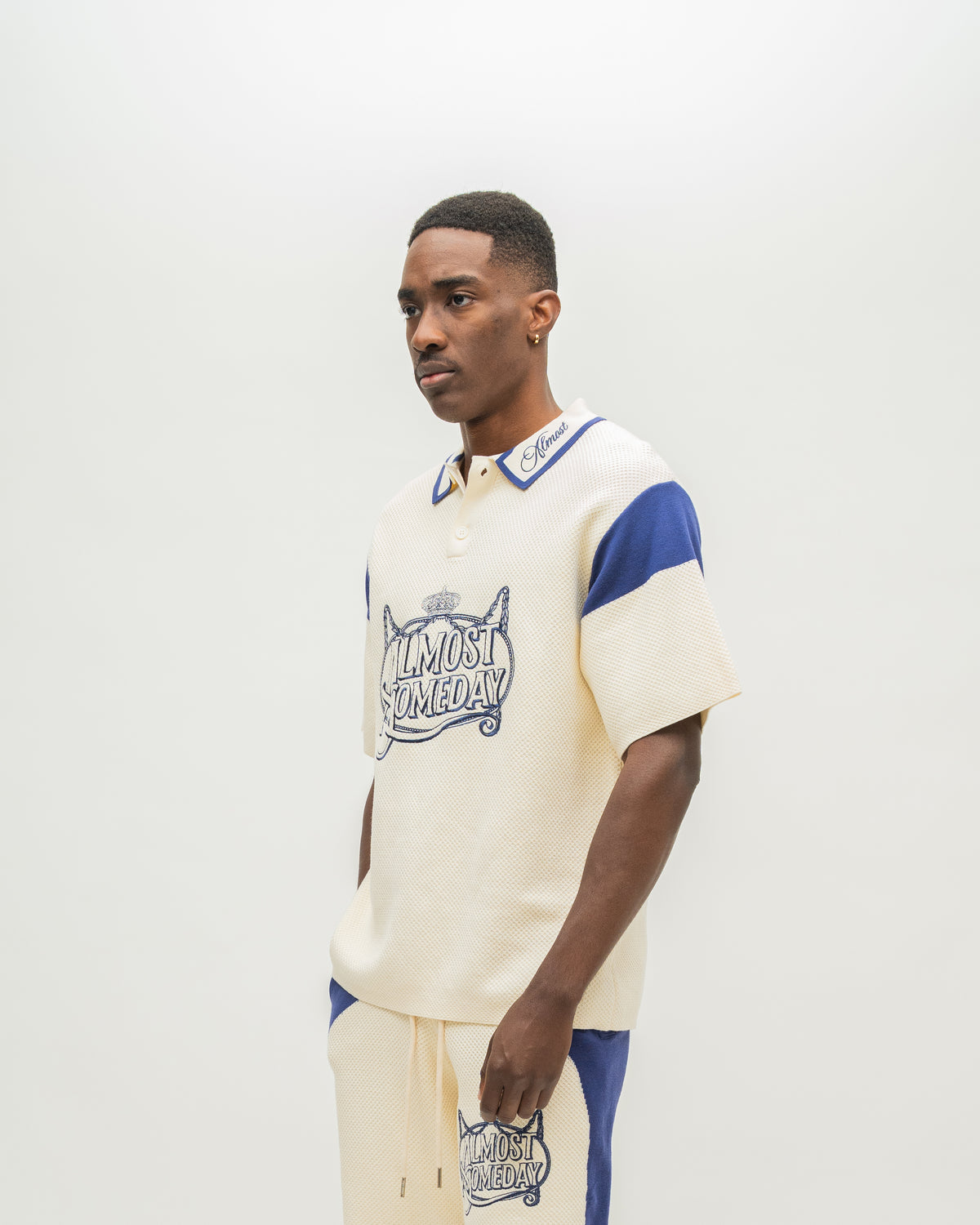 A man modeling a cream-colored short-sleeve polo shirt with blue accents and a graphic print on the front, paired with matching cream pants with blue side panels, against a plain light gray background.