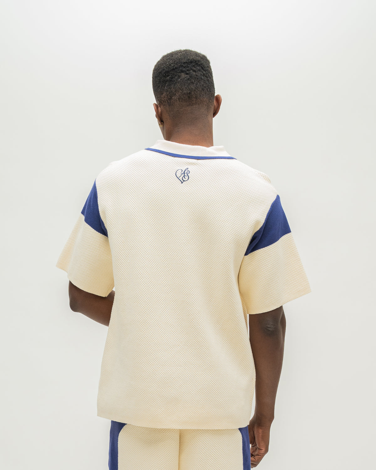 A man viewed from behind wearing a cream-colored short-sleeve knit top with blue shoulder panels and a small blue chest logo, paired with matching cream and blue shorts, against a plain white background.