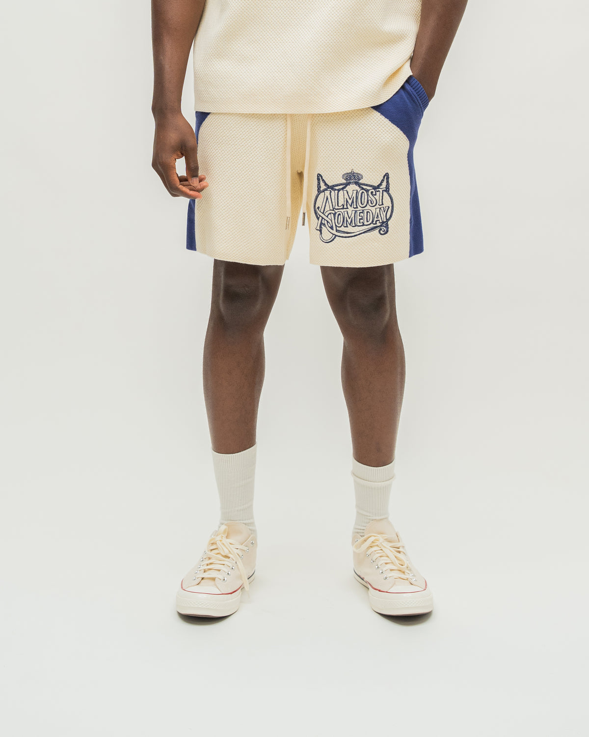 A model is shown from the waist down wearing cream-colored shorts with blue side panels and a navy graphic print on the left leg. The shorts feature a drawstring waist and are paired with cream socks and cream sneakers on a light gray studio background.