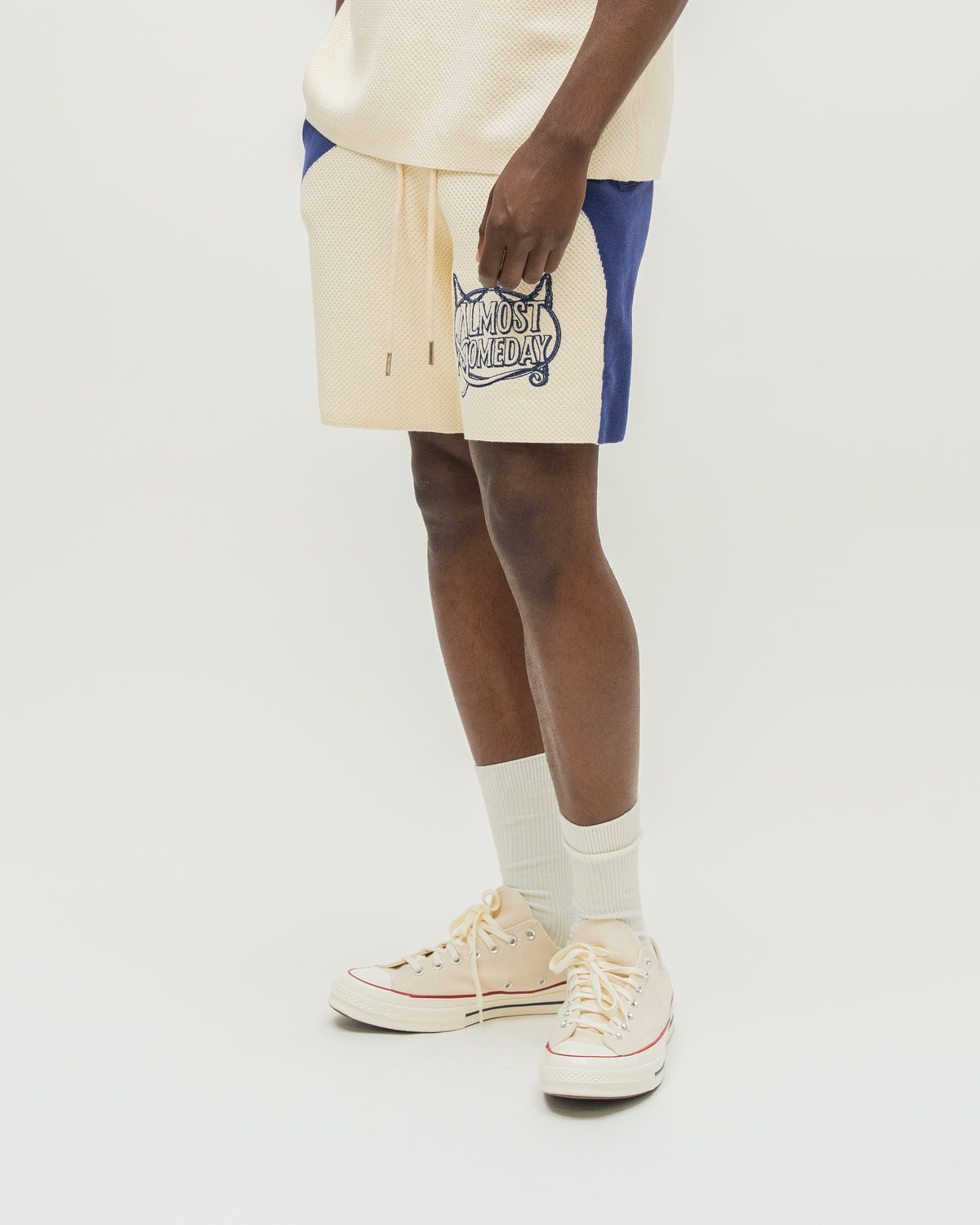 A person wearing cream shorts with blue side panels and a blue graphic print, paired with cream socks and cream sneakers, against a plain light gray studio background.