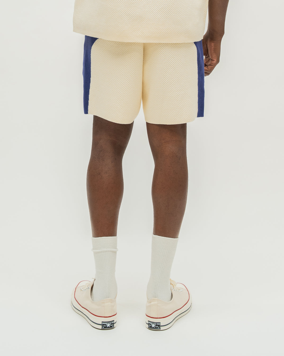 A person shown from the back wearing cream knit shorts with blue side panels, white socks, and cream low-top sneakers against a plain light background.
