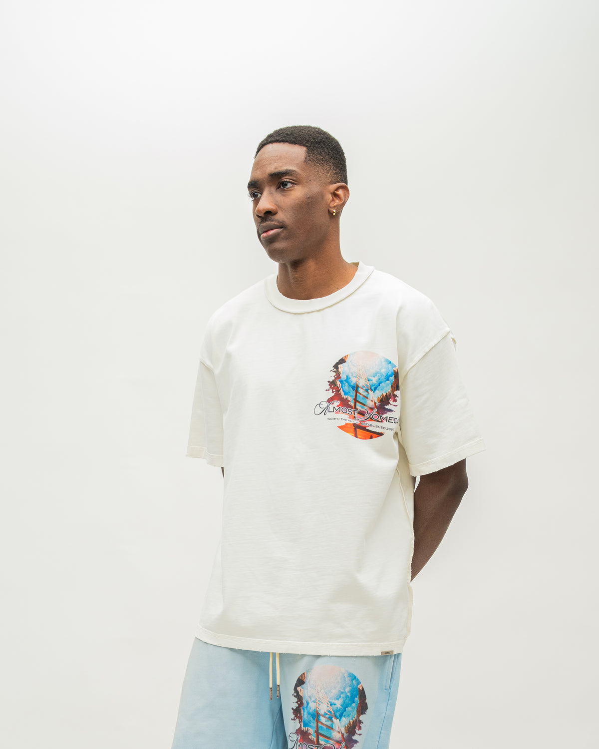 A man wearing a cream-colored oversized t-shirt with a colorful graphic print on the left chest, paired with light blue sweatshorts, standing against a plain off-white studio background.