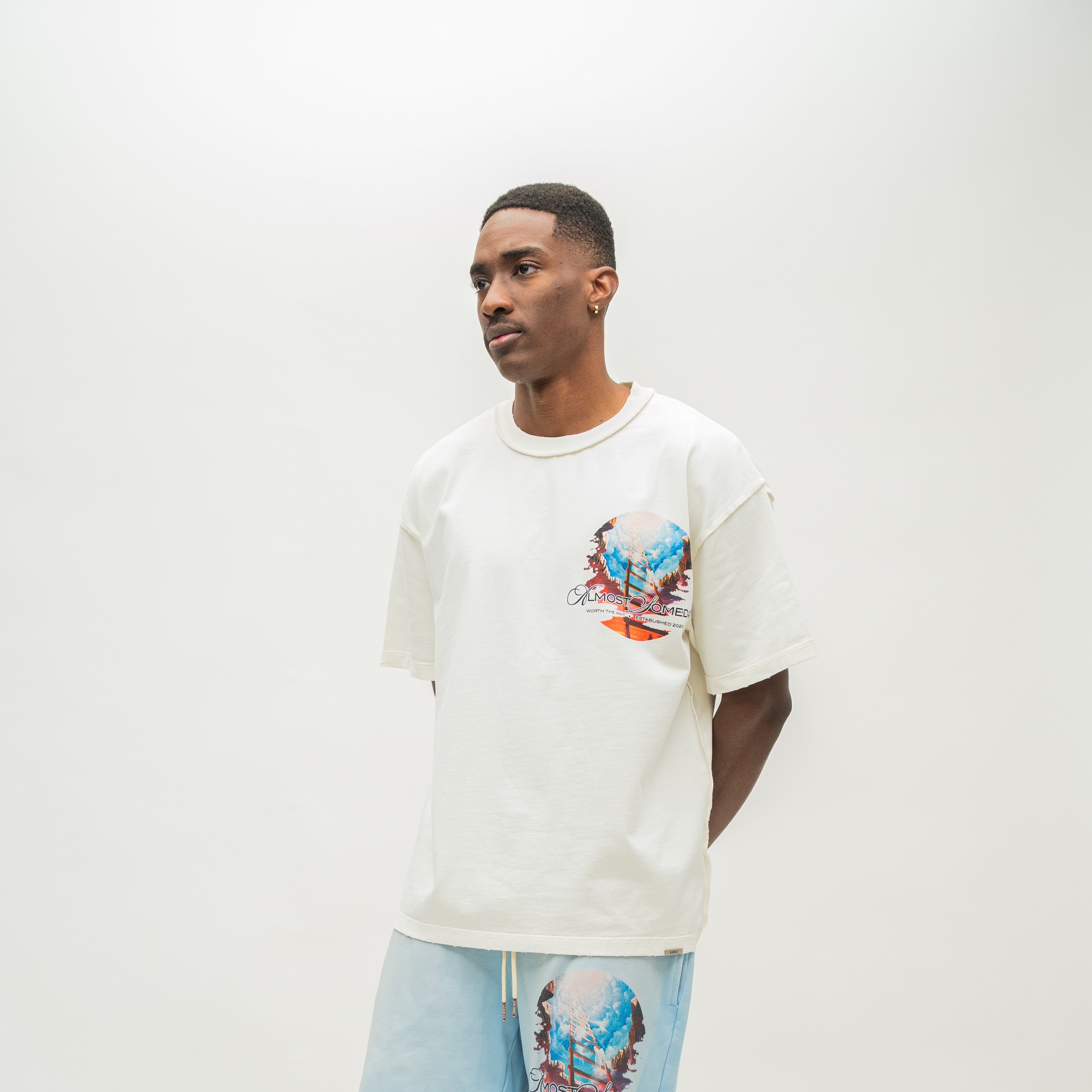 A man wearing a cream-colored oversized t-shirt with a colorful graphic print on the left chest, paired with light blue sweatshorts, standing against a plain off-white studio background.