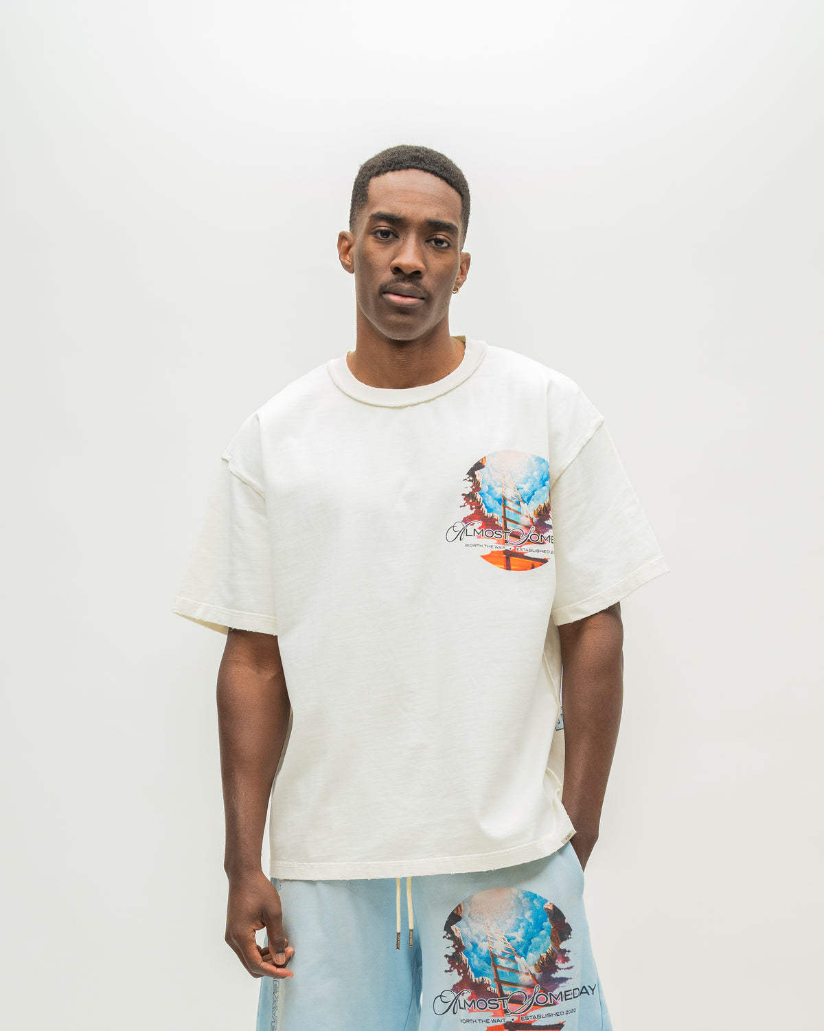A man wearing an oversized off-white graphic t-shirt and light blue shorts with a matching graphic print, standing against a plain light gray studio background.
