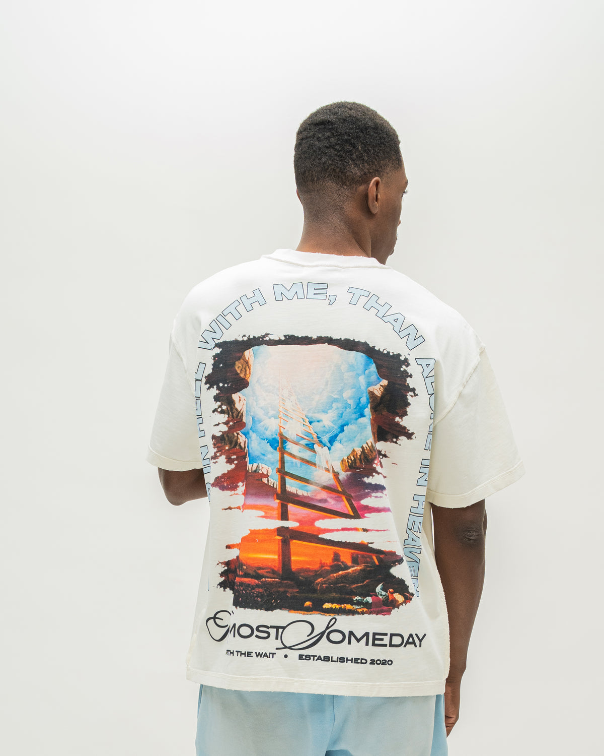 A man viewed from behind wearing a white Almost Someday graphic t-shirt with a large colorful print of a sailing ship and text, paired with light blue sweatshorts against a plain white background.