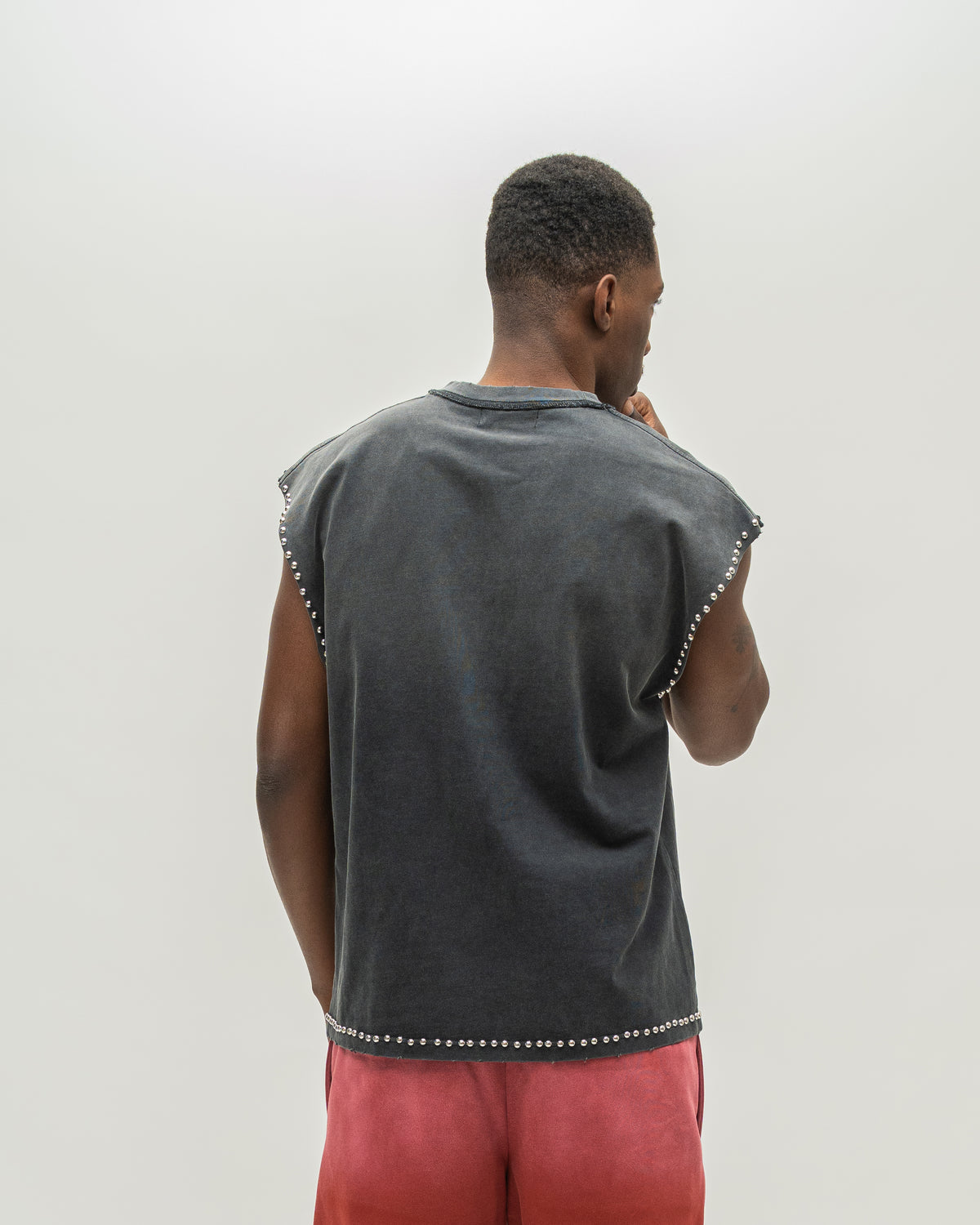 A man viewed from behind wearing a dark gray sleeveless t-shirt with silver stud detailing along the edges, paired with coral pink pants, against a plain light gray background.