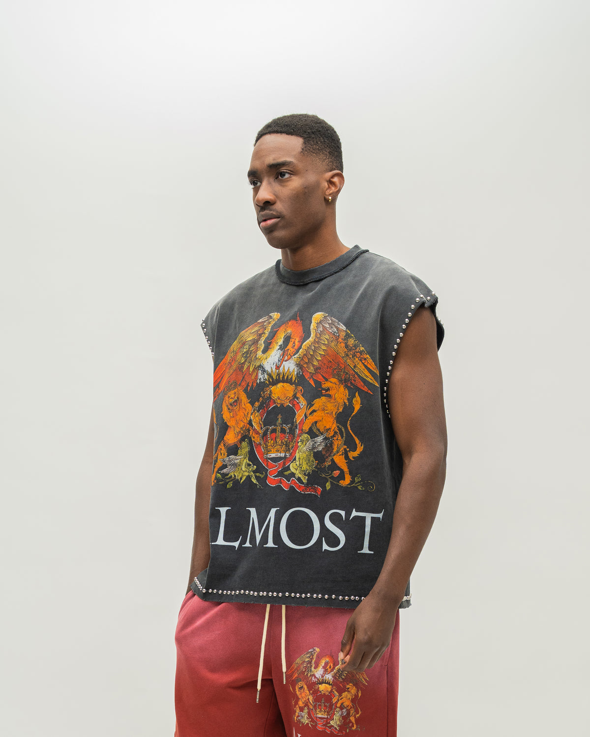 A man modeling a sleeveless black t-shirt with a large orange and gold eagle graphic and the word 'ALMOST' printed on the front, paired with red sweatpants against a plain light gray background.