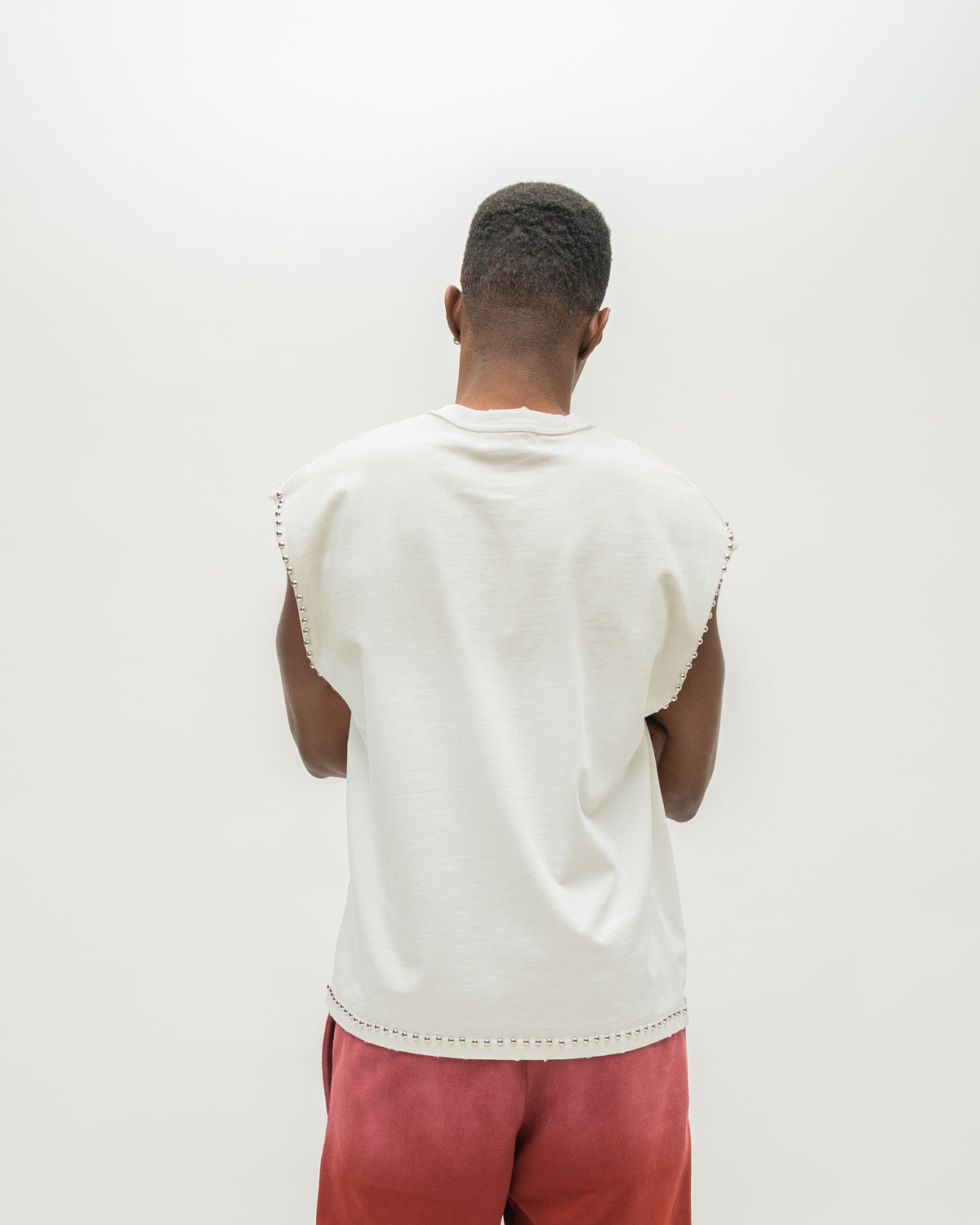 A man viewed from behind wearing a sleeveless white t-shirt with scalloped edges and pink pants, standing against a plain white background.