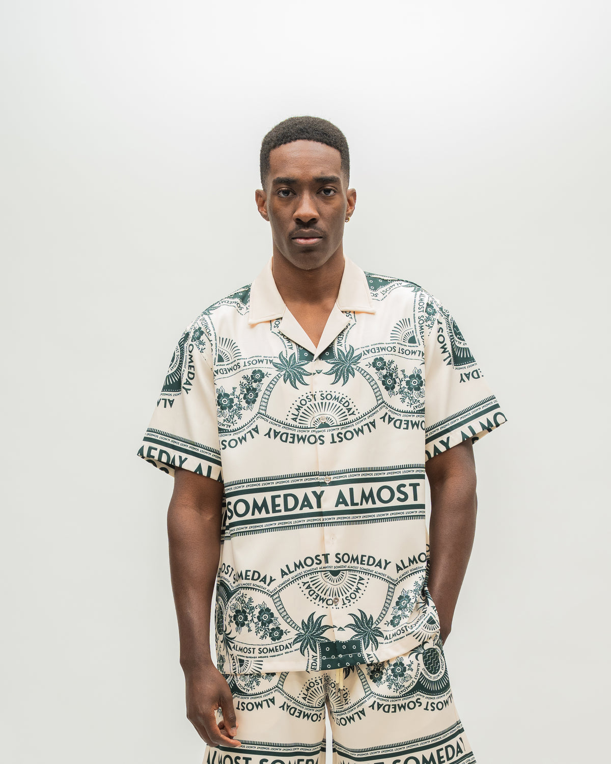 A man wearing a matching short-sleeve camp collar shirt and shorts set with a cream base and dark green graphic print featuring palm trees and the text 'ALMOST SOMEDAY'.