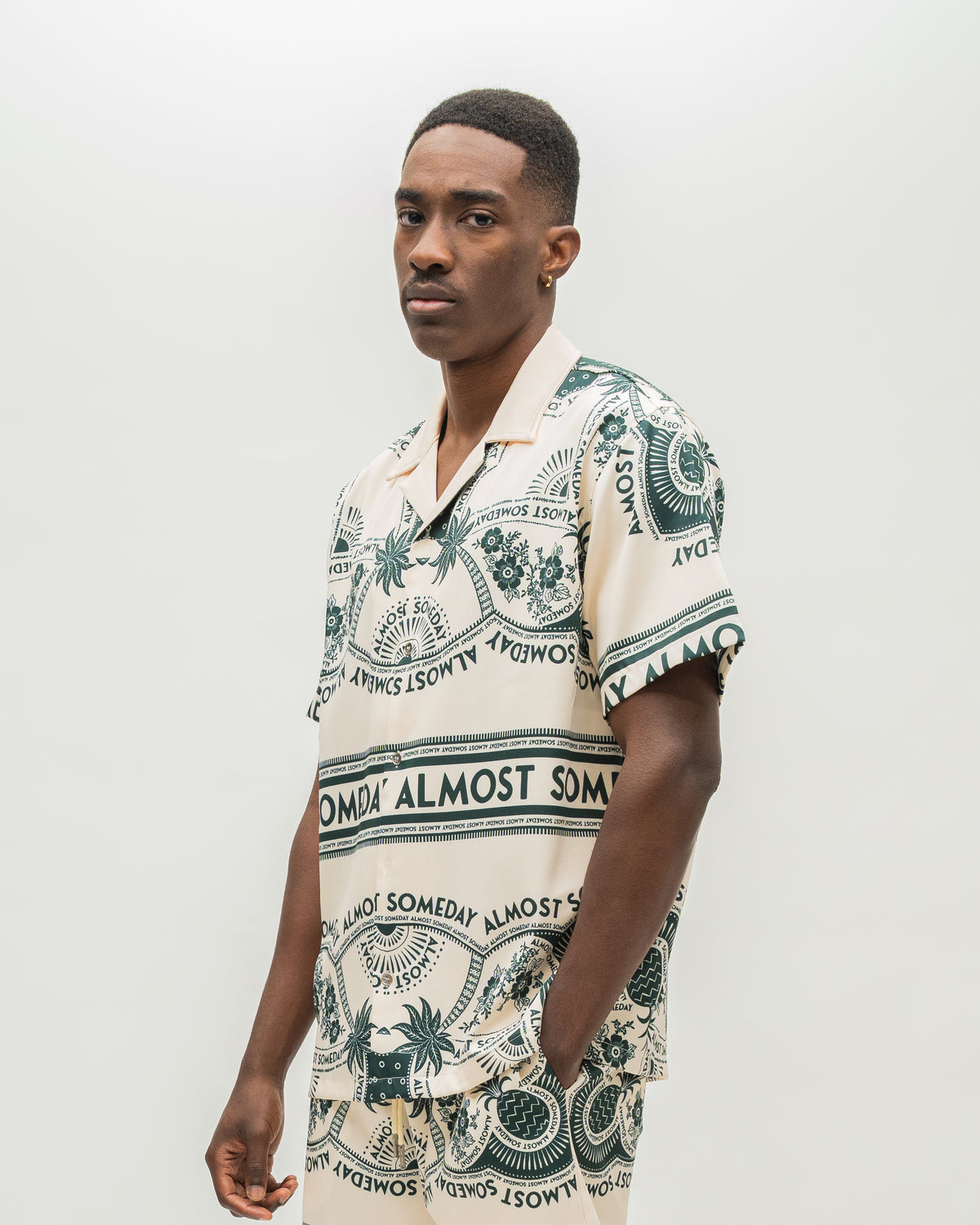 A man wearing a short-sleeve camp collar shirt with a cream base and dark green tropical-themed print featuring palm trees, pineapples, and the text 'ALMOST SOMEDAY'.