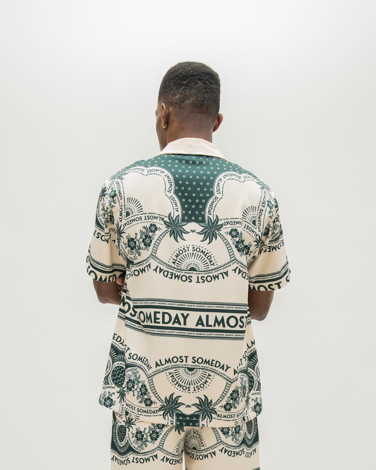 A man viewed from behind wearing a matching short-sleeve button-up shirt and shorts set in cream with a dark green graphic print featuring palm trees and the text 'ALMOST SOMEDAY'.