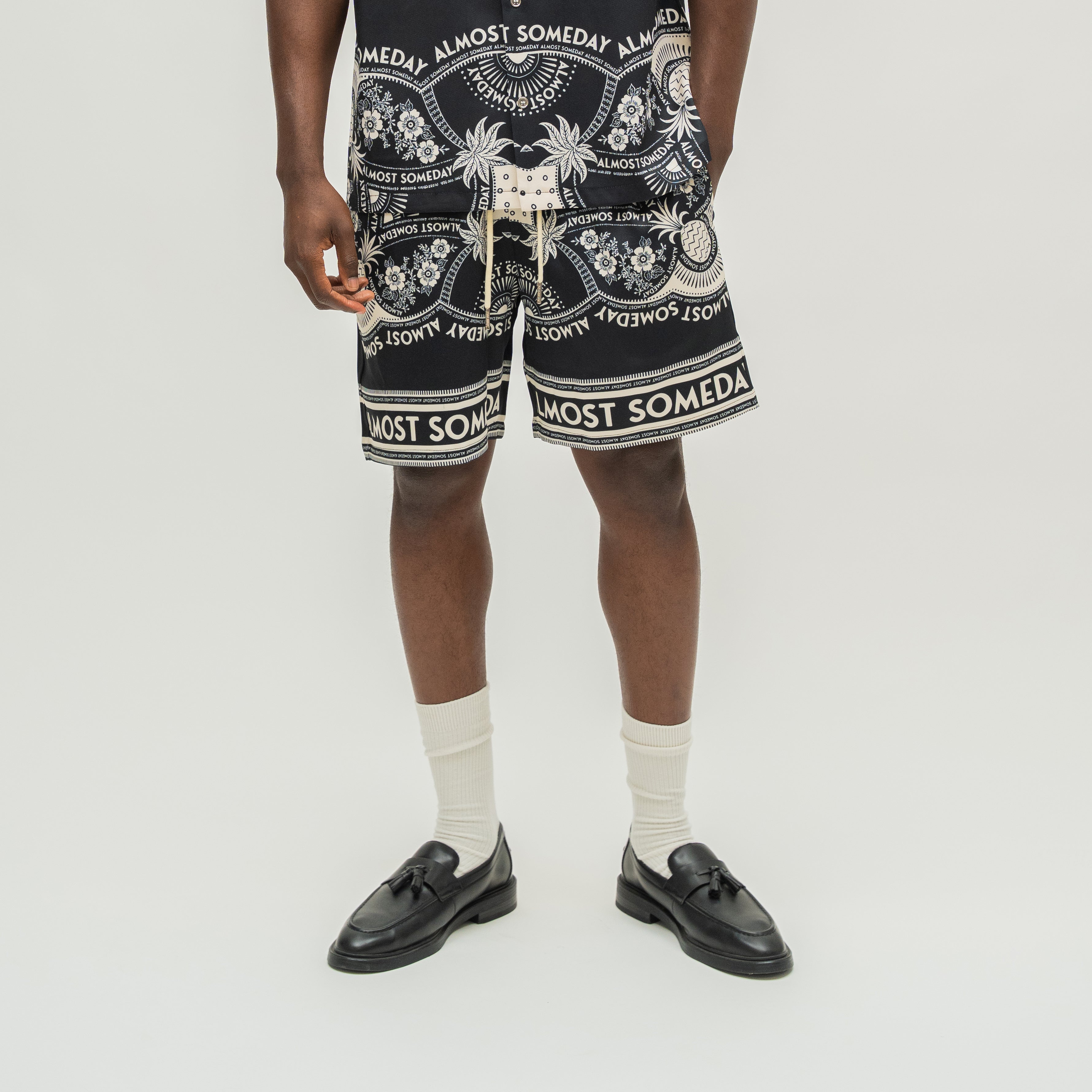 A model from the waist down wearing black and white patterned drawstring shorts, white socks, and black leather loafers, standing against a light gray studio background.