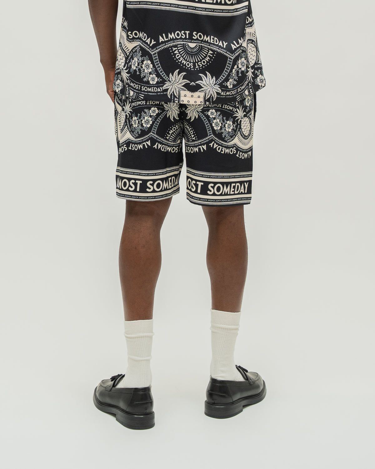 A person shown from the back wearing black shorts with a white ornate graphic print and text, paired with white socks and black loafers, against a light gray studio background.