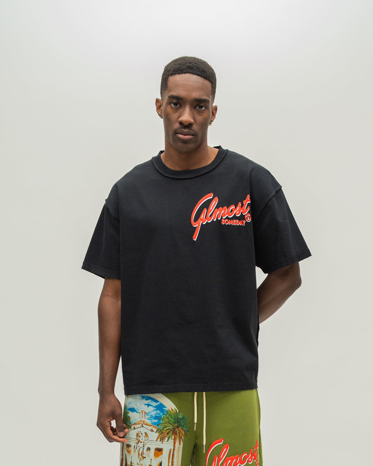 A man wearing a black oversized t-shirt with a red 'Almost Someday' logo on the chest, paired with olive green shorts featuring a tropical beach graphic and red lettering, set against a plain light gray background.