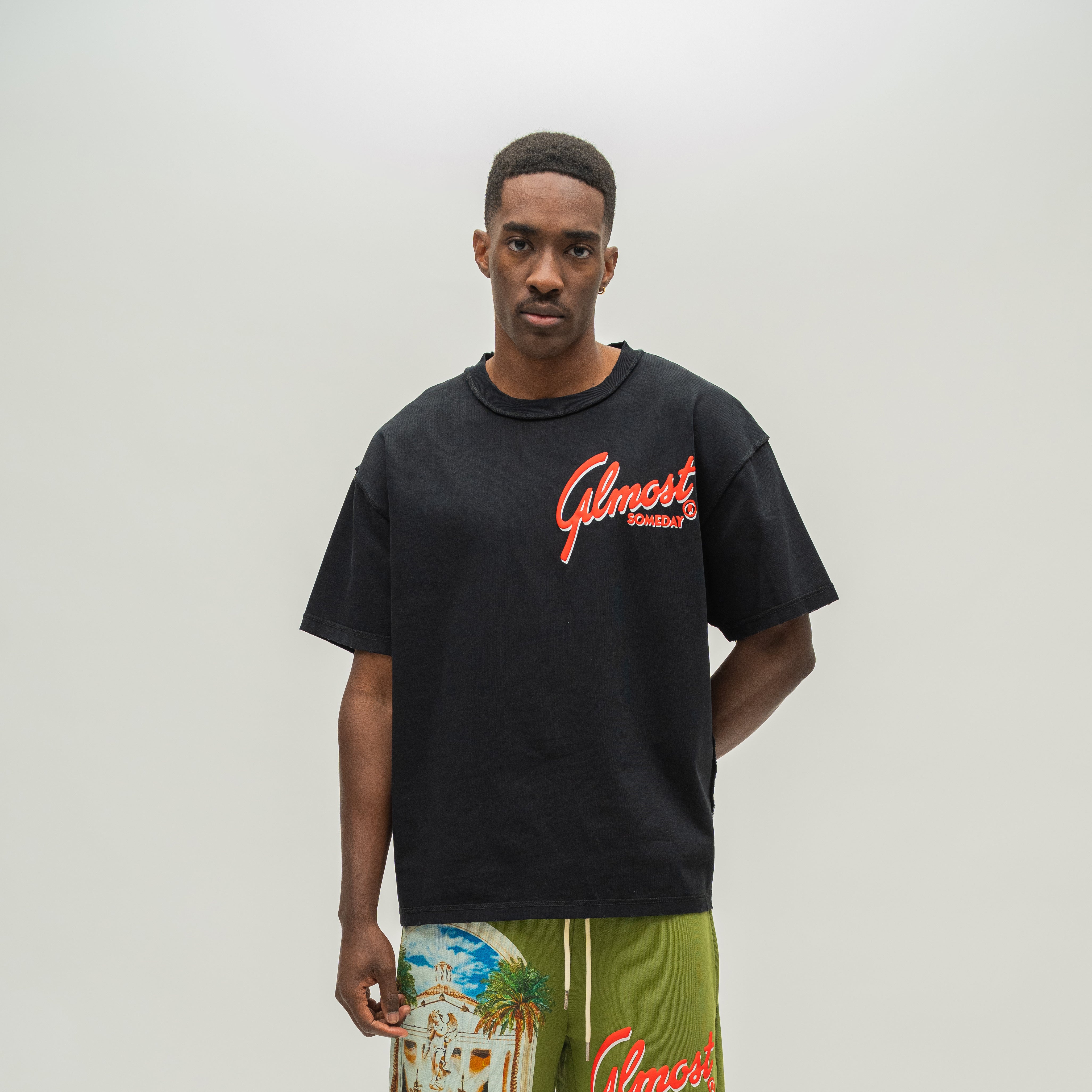 A man wearing a black oversized t-shirt with a red 'Almost Someday' logo on the chest, paired with olive green shorts featuring a tropical beach graphic and red lettering, set against a plain light gray background.