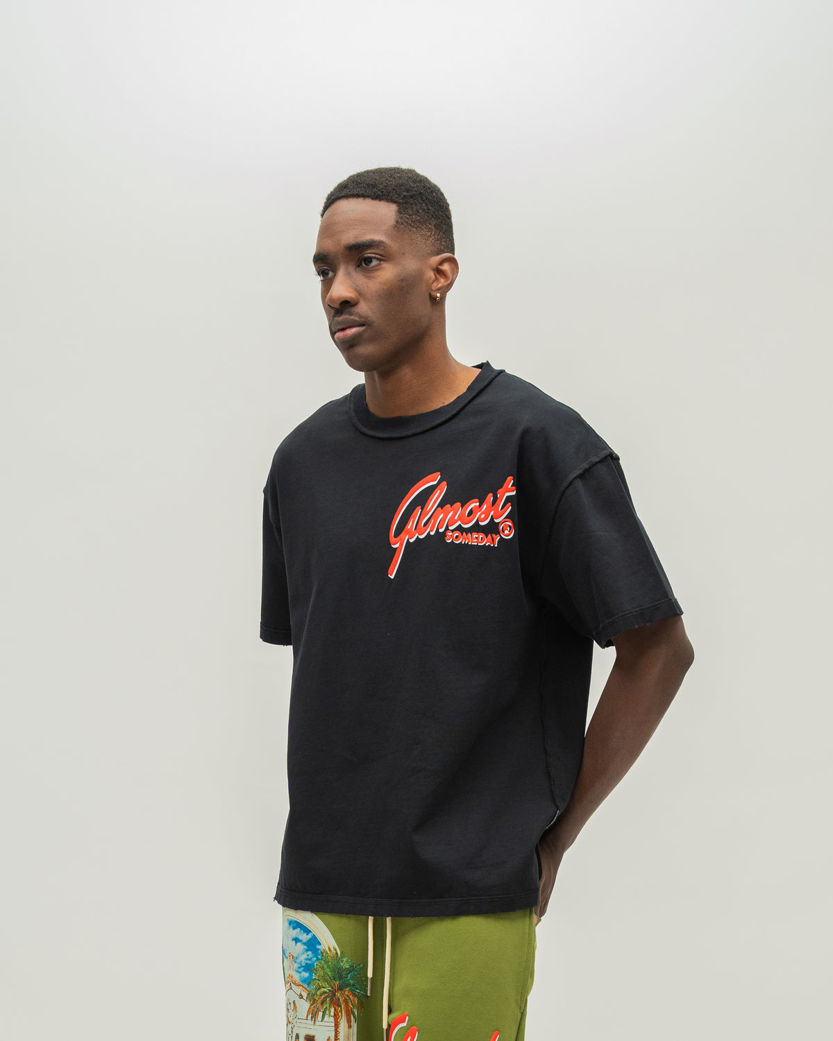 A man wearing a black crewneck t-shirt with a red and orange 'Almost Someday' graphic on the chest, paired with olive green tropical print shorts, standing against a plain light gray background.