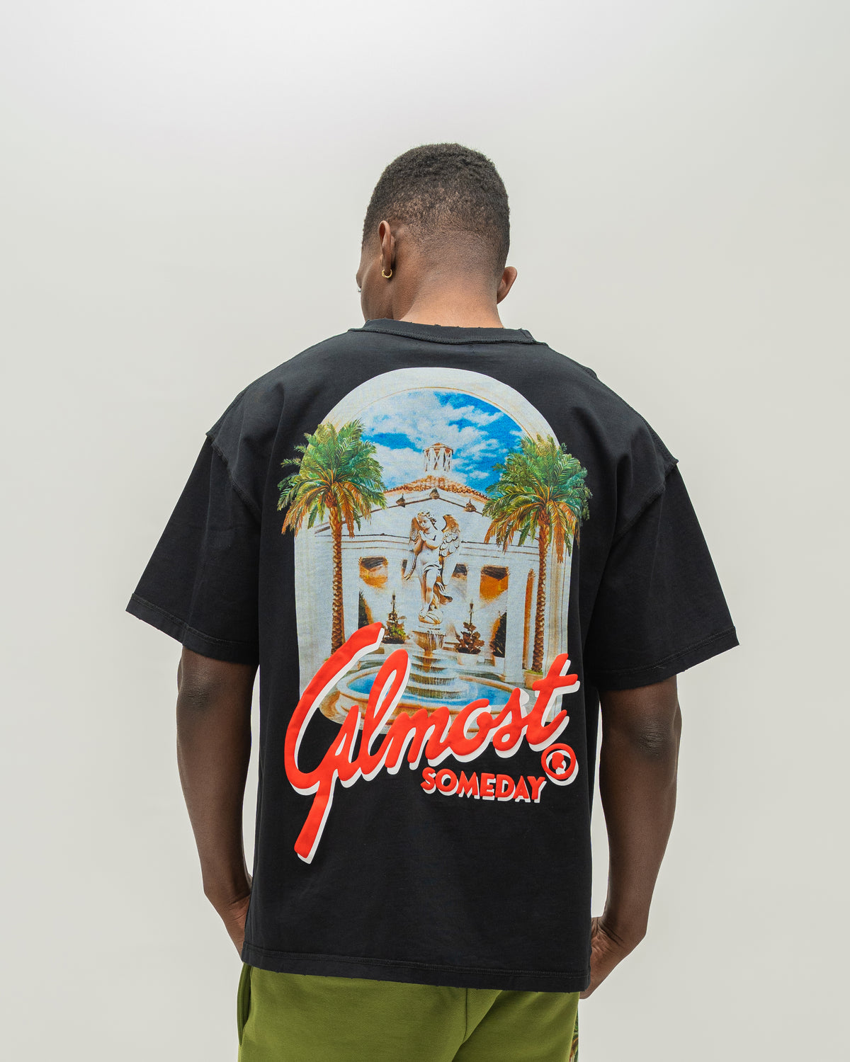 A man viewed from behind wearing a black t-shirt with a large graphic print on the back. The graphic features a white building with palm trees under a blue sky, with the red text 'ALMOST SOMEDAY' written across the bottom.