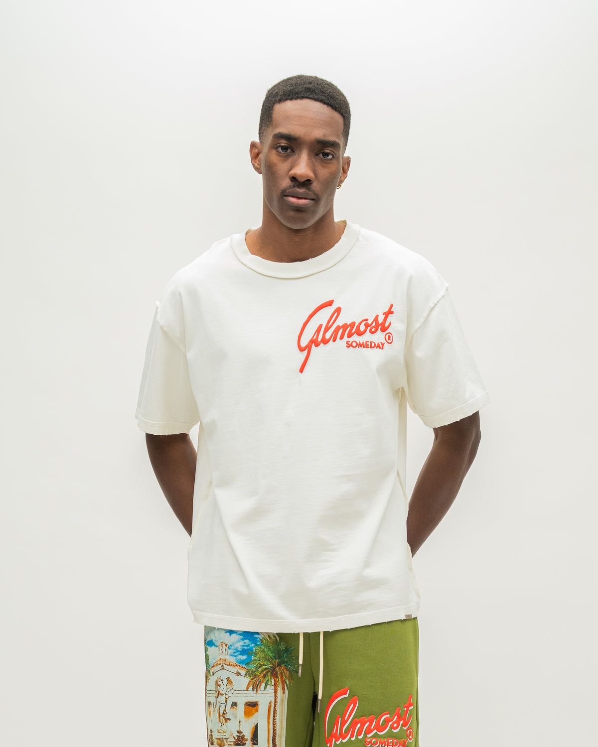 A man wearing a cream-colored t-shirt with red 'Almost Someday' script on the chest, paired with olive green shorts featuring a tropical beach print and the 'Almost Someday' logo. The background is a clean, off-white studio wall.