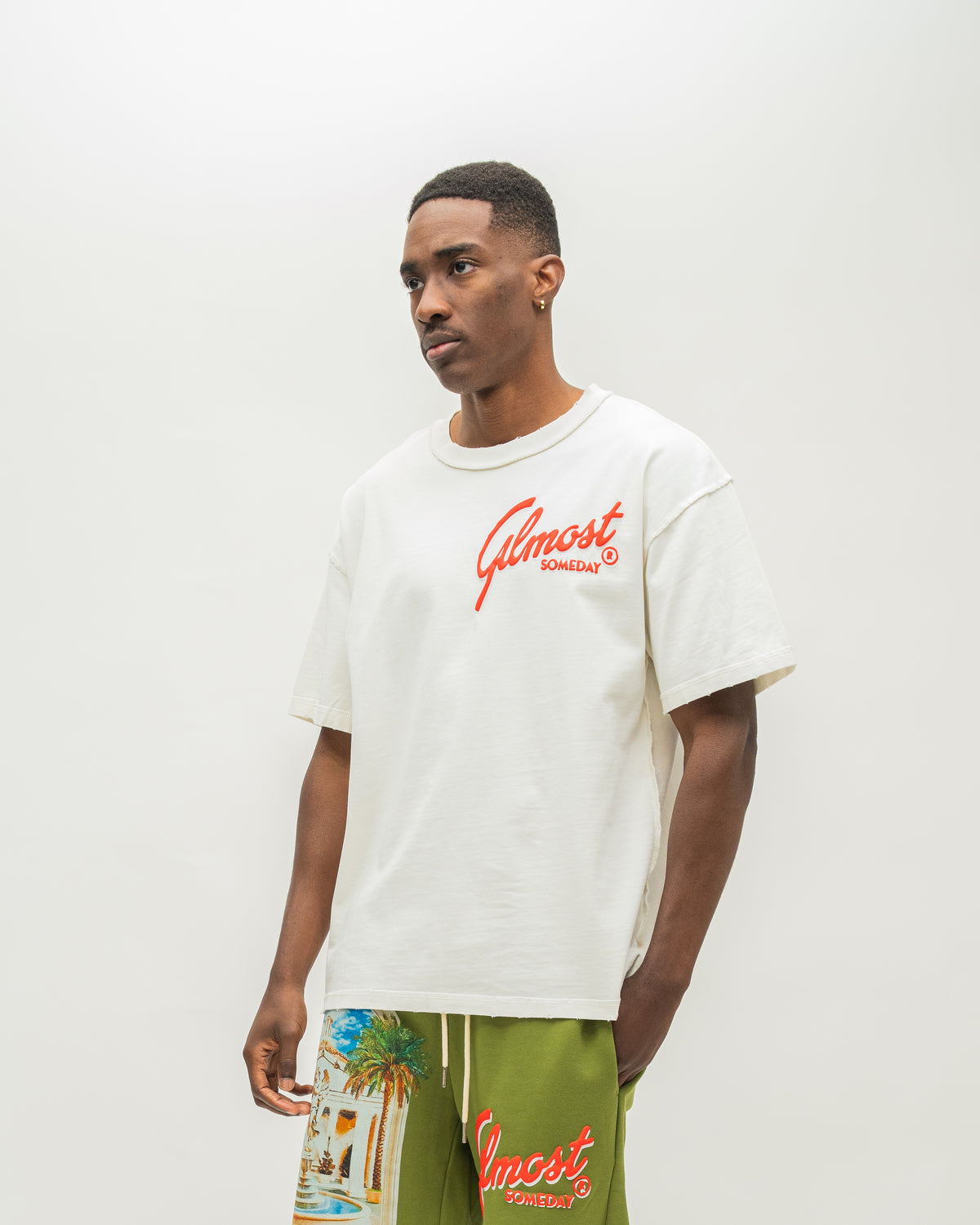 A man wearing an off-white t-shirt with a red 'Almost Someday' logo and green shorts with a beach graphic and red logo, standing against a plain light gray background.