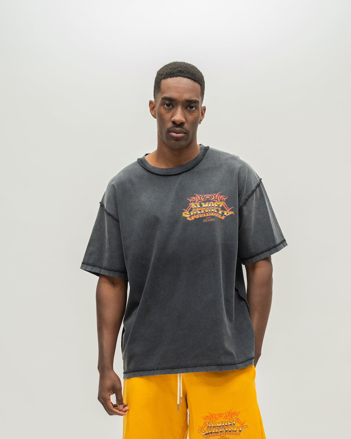 A man modeling an oversized faded black acid-wash t-shirt with a distressed orange graphic on the left chest, paired with bright yellow sweatpants, against a plain light gray background.