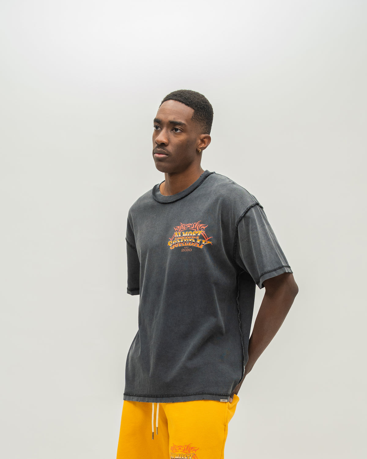 A man wearing a washed black t-shirt with a yellow and red graphic on the chest, paired with bright yellow sweatshorts, standing against a light gray studio background.