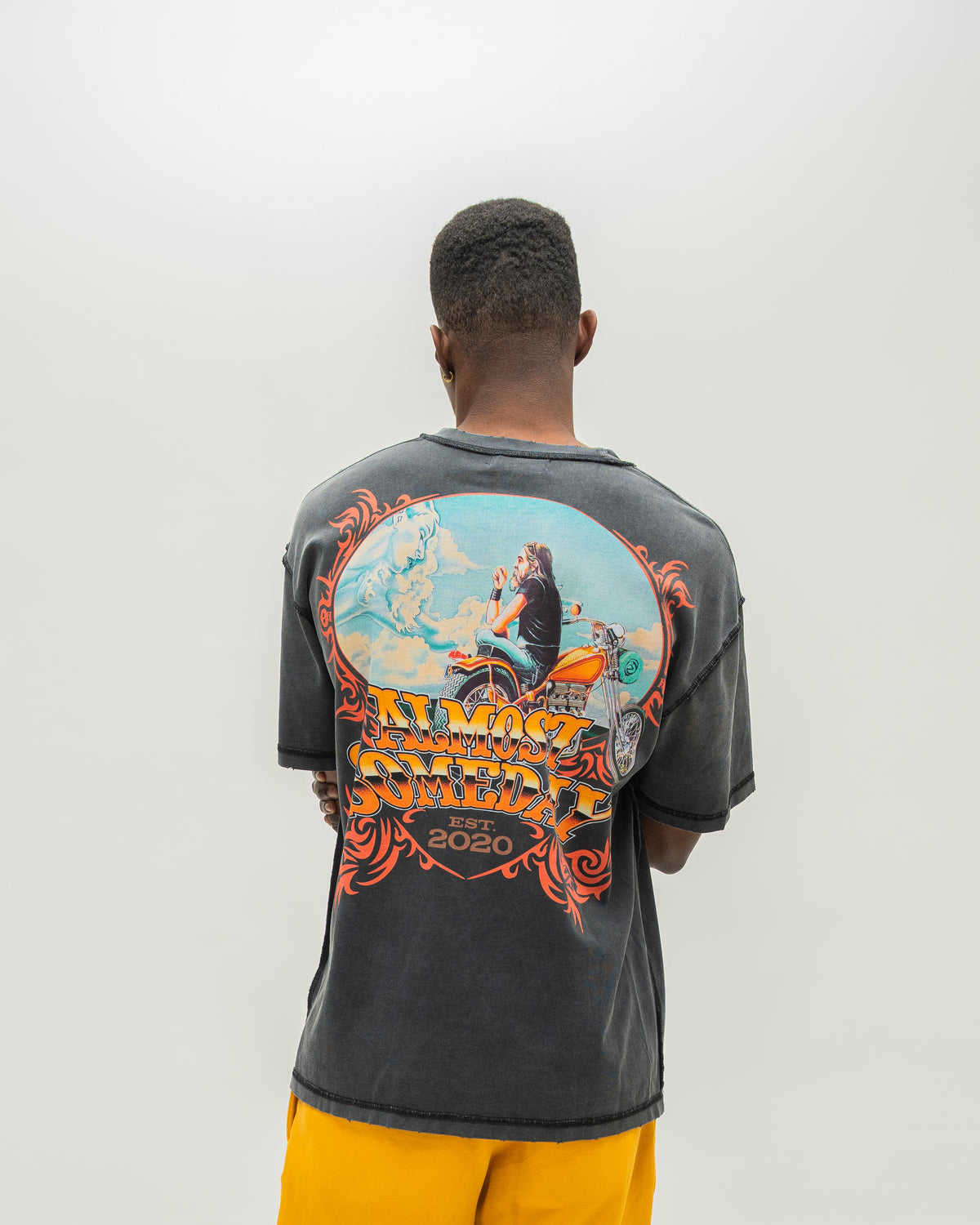 A man viewed from behind wearing a faded black mineral-washed t-shirt with a large colorful graphic on the back, paired with yellow sweatshorts, against a plain light gray background.