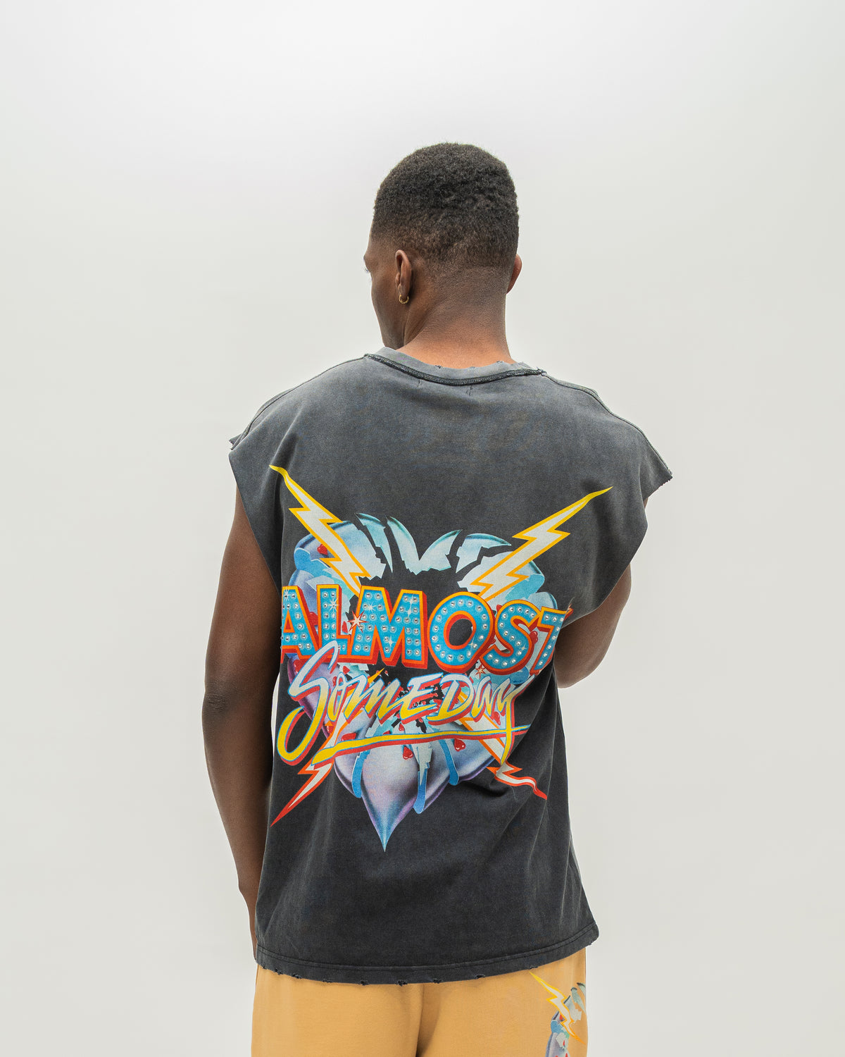 A man viewed from behind wearing a faded black sleeveless t-shirt with a large colorful graphic on the back reading 'ALMOST SOMEDAY', paired with yellow shorts, against a plain light gray background.