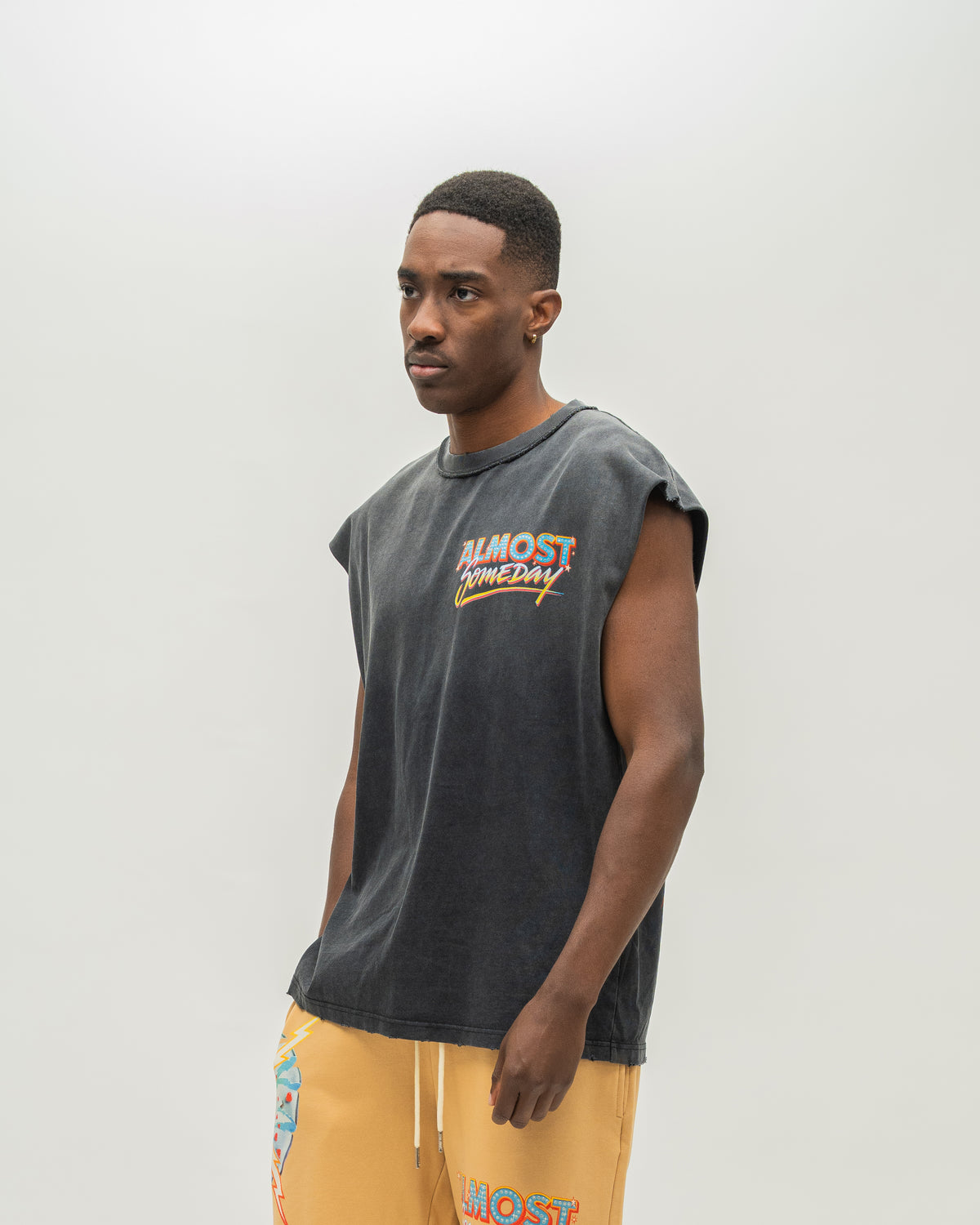 A man wearing a faded black sleeveless t-shirt with a colorful 'ALMOST SOMEDAY' graphic on the chest, paired with yellow sweatshorts, standing against a light gray studio background.