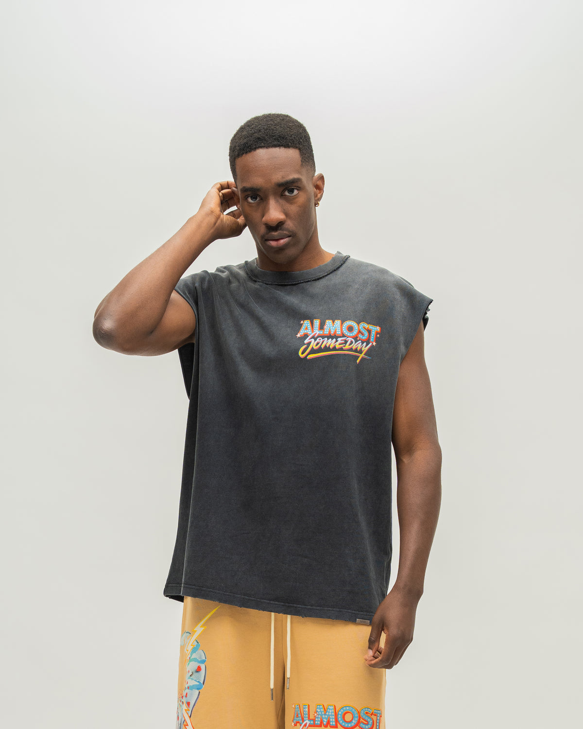 A man modeling a faded black sleeveless t-shirt with a distressed graphic that reads 'ALMOST SOMEDAY' in orange and yellow, paired with yellow drawstring shorts against a light gray studio background.