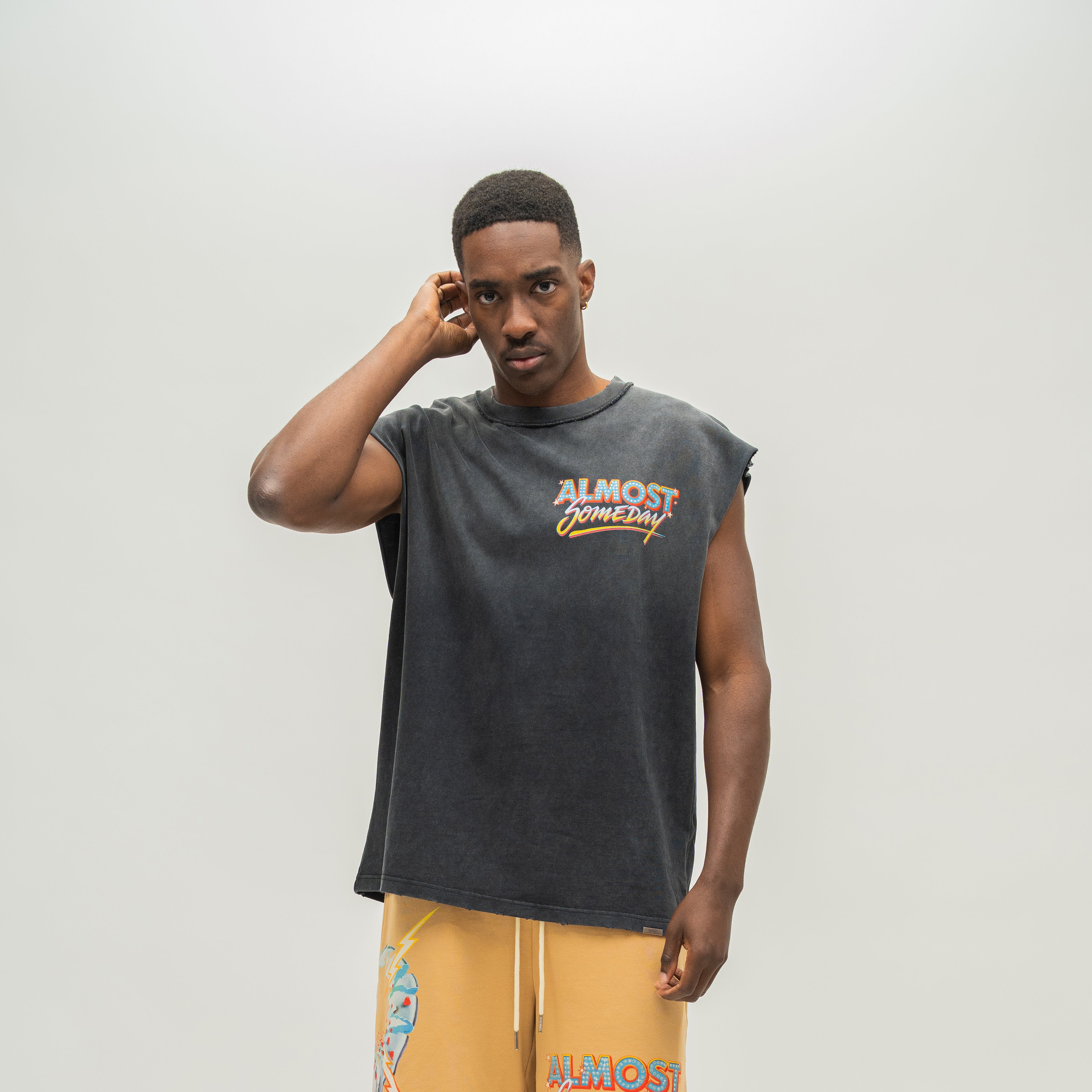 A man modeling a faded black sleeveless t-shirt with a distressed graphic that reads 'ALMOST SOMEDAY' in orange and yellow, paired with yellow drawstring shorts against a light gray studio background.