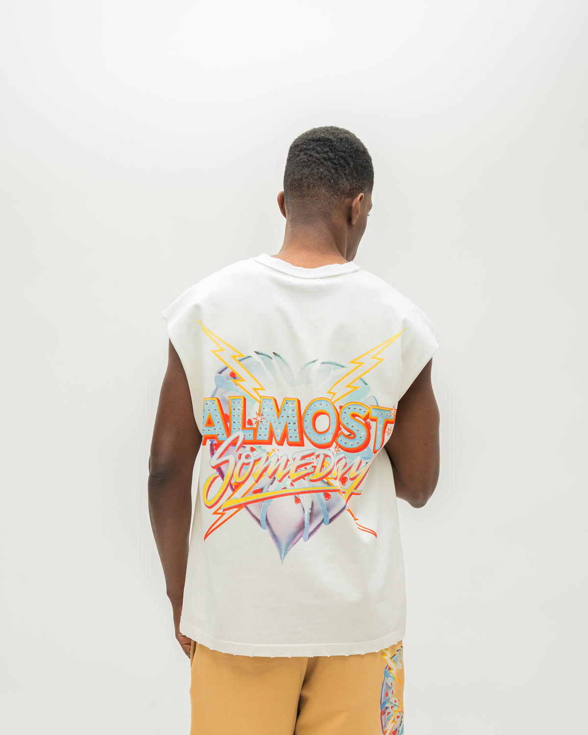 A man viewed from behind wearing a sleeveless white t-shirt with a colorful 'ALMOST SOMEDAY' graphic on the back, paired with yellow shorts, against a plain white studio background.