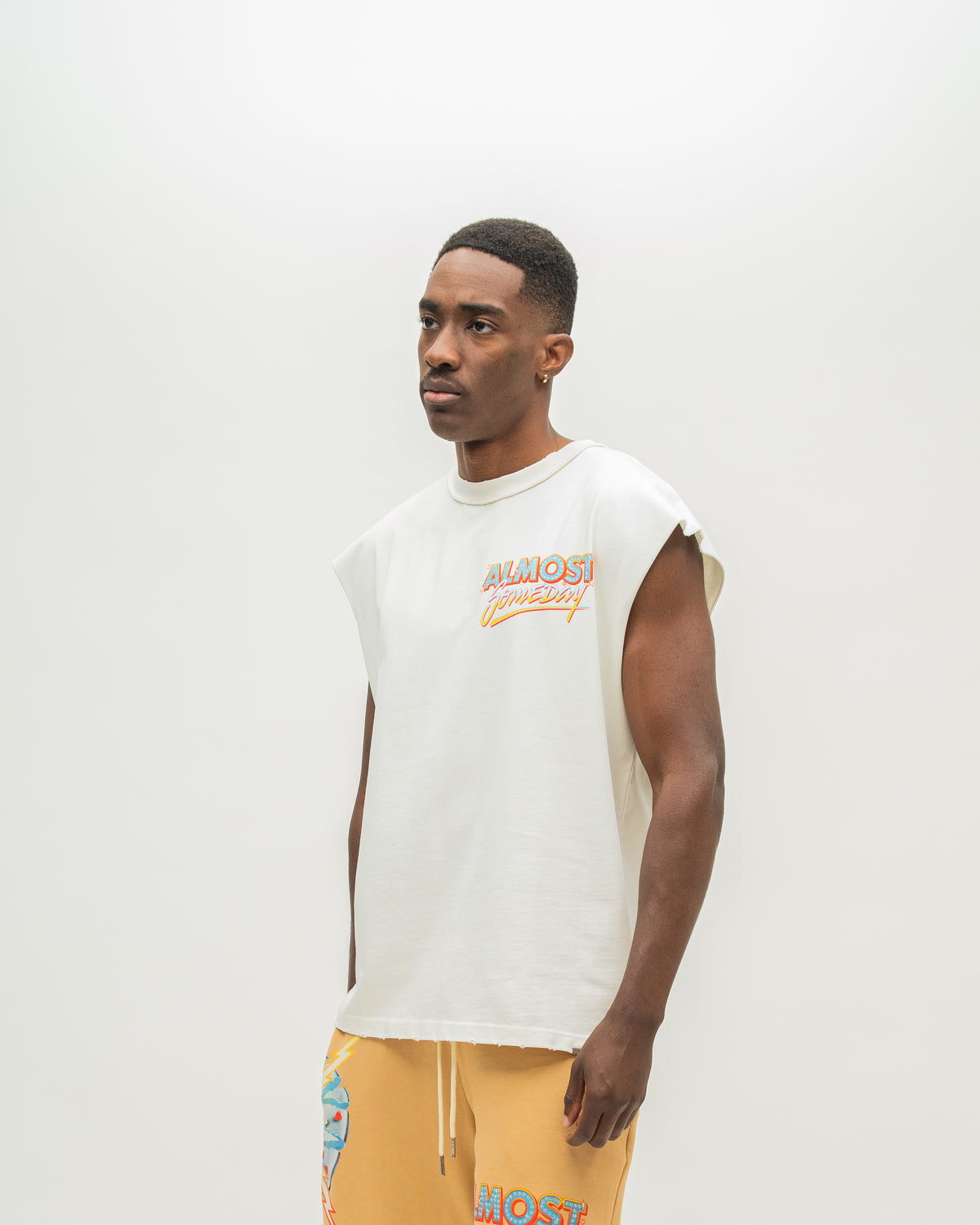 A man wearing a sleeveless white t-shirt with a colorful 'ALMOST SOMEDAY' graphic on the chest, paired with mustard yellow sweatshorts, photographed against a plain light gray studio background.