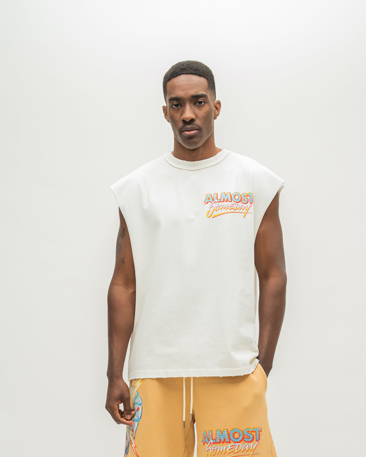 A man modeling a sleeveless white t-shirt with a colorful 'ALMOST SOMEDAY' logo on the chest, paired with mustard yellow sweatshorts against a plain light gray background.