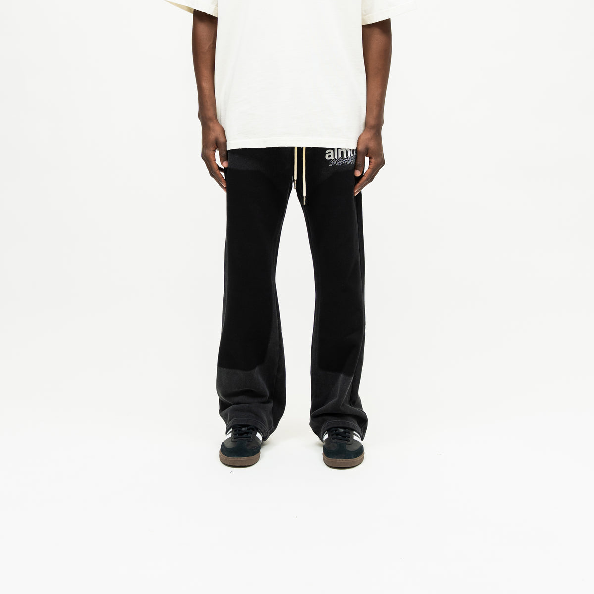 Enemies Flare Bottoms – Black – Front view on model styled with a cream tee, showing white drawstrings, thigh logo, and flared legs