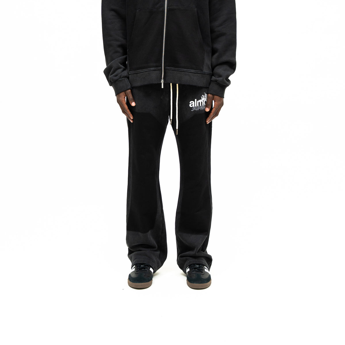 Enemies Flare Bottoms – Black – Front view on model styled with a black zip hoodie, showing white drawstrings and thigh logo