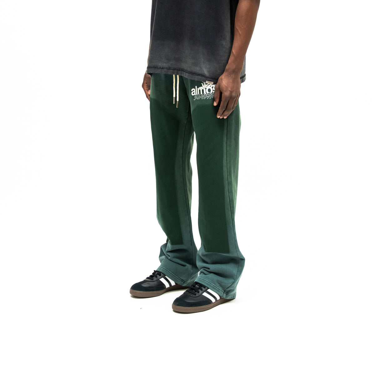 Enemies Flare Bottoms – Green – Three-quarter front view on model styled with a black tee, showing thigh logo, dark leg panels, and flared hems