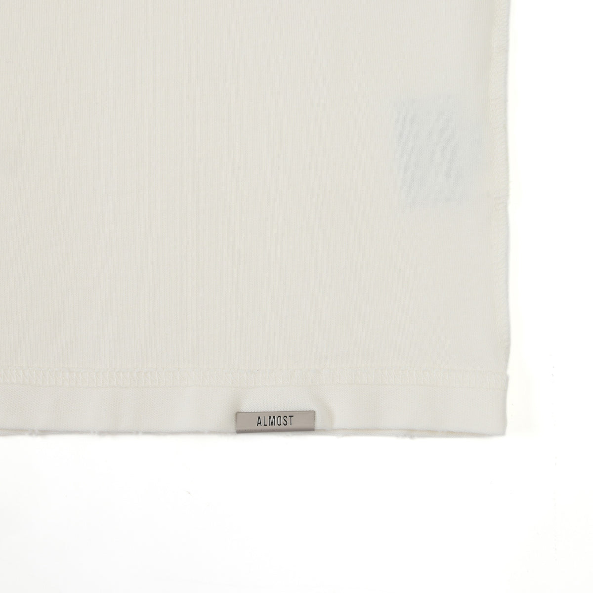 Enemies Tee – Cream – Close-up of bottom hem with metal ALMOST logo tab