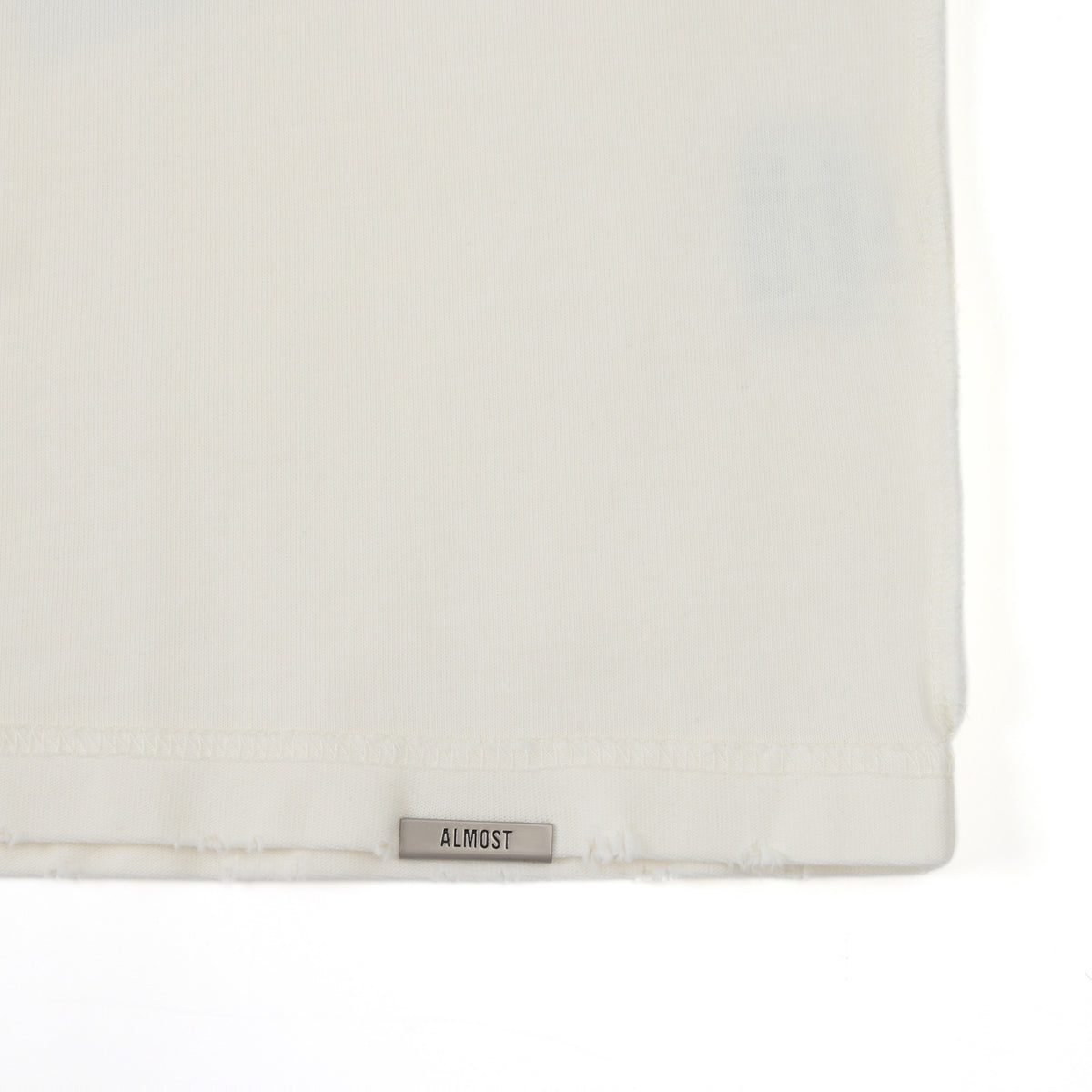 Members Tee – Cream – Close-up of metal ALMOST logo at the bottom hem