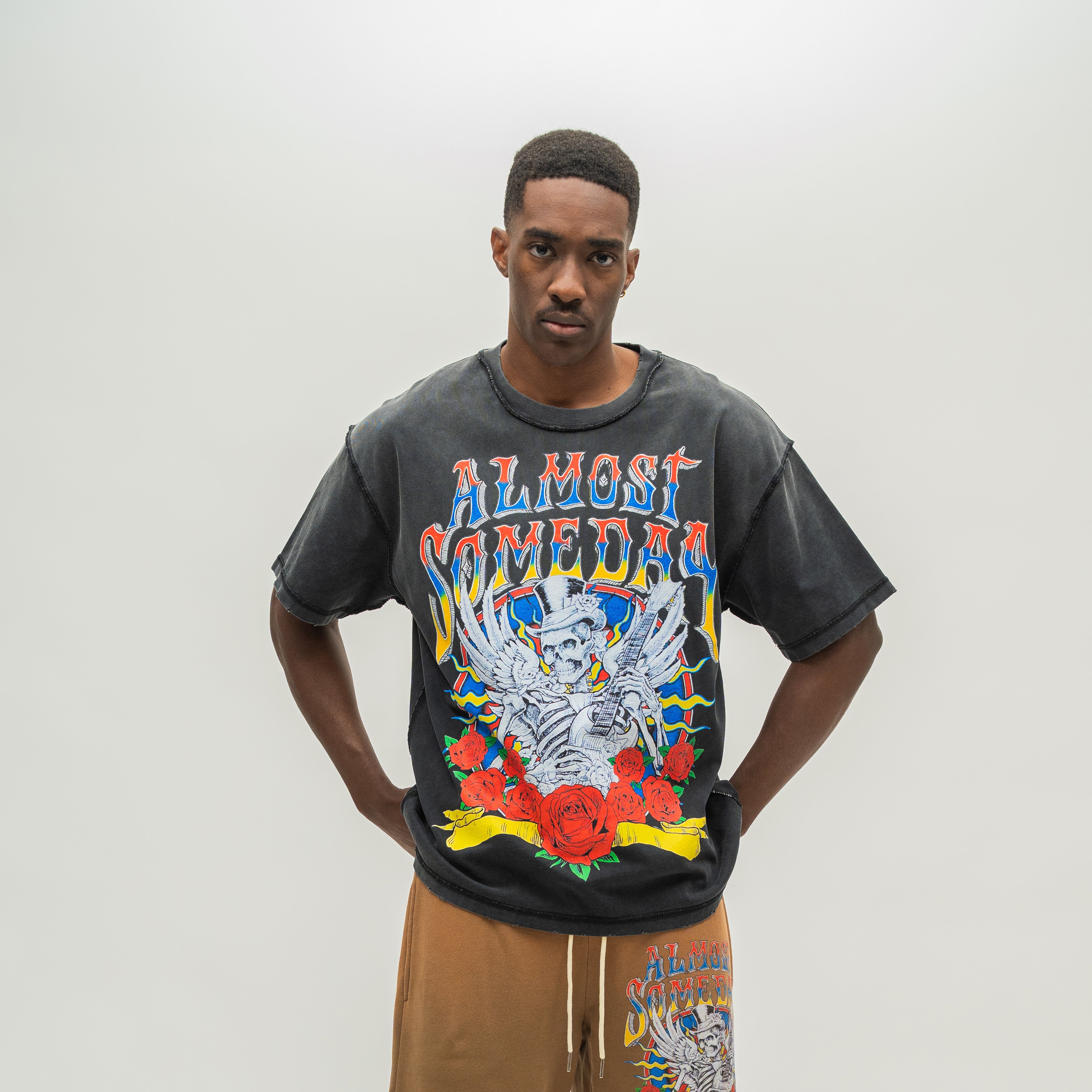 A man modeling a washed black graphic t-shirt with a colorful 'ALMOST SOMEDAY' band logo and skeleton/rose design, paired with brown drawstring shorts against a plain light gray background.